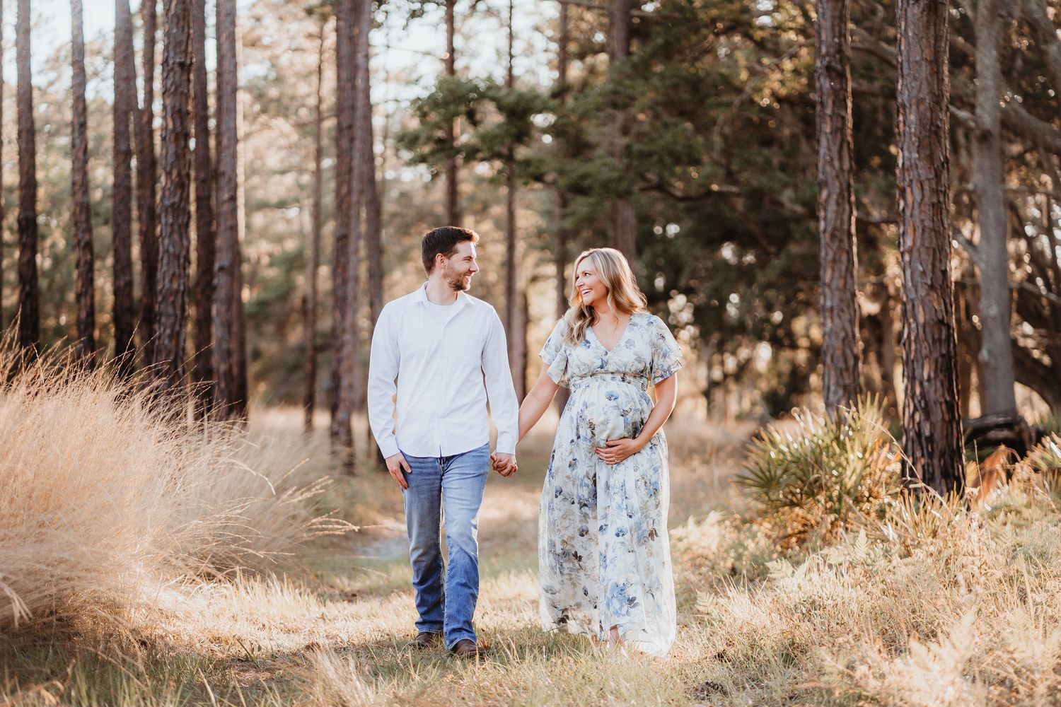 Smiling pregnant couple strolling through golden meadow surrounded by pine trees.