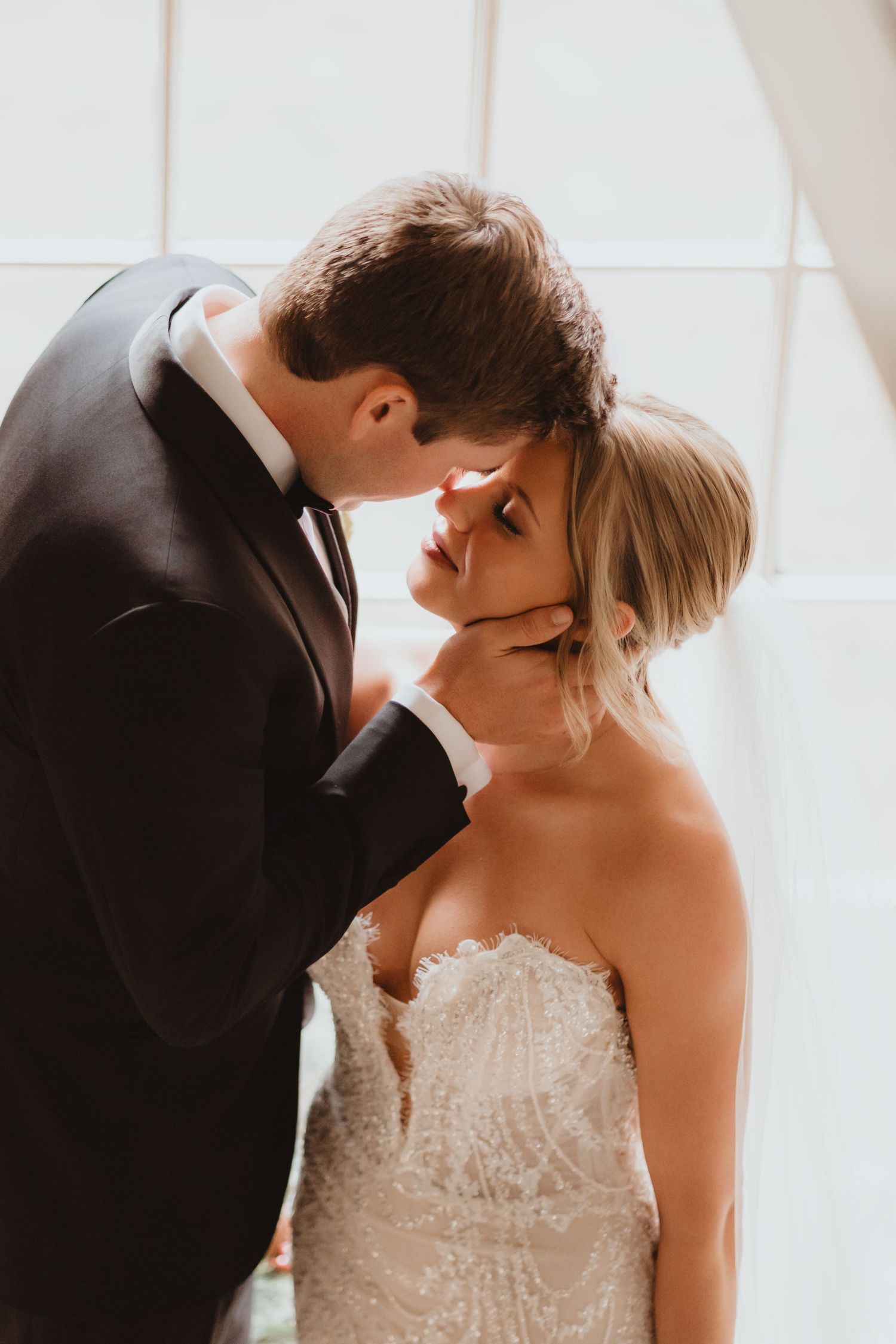 Groom gently cups brides face for an intimate moment in soft window light.