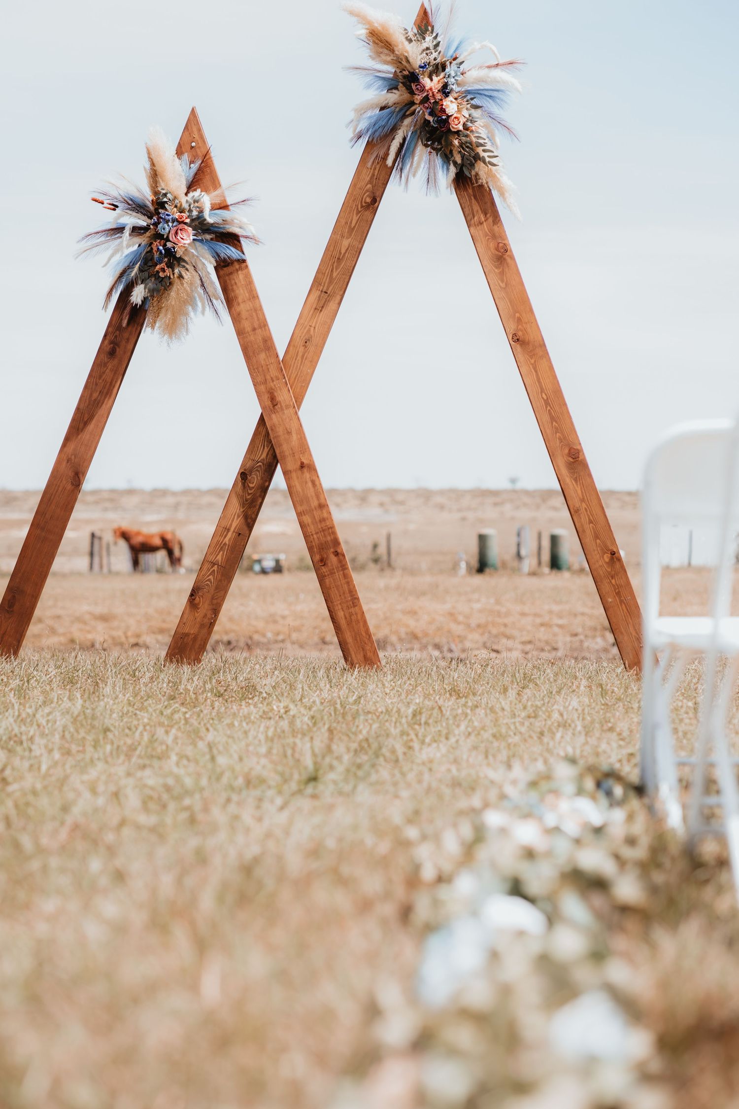 Two wooden triangular wedding arches decorated with boho floral arrangements in open field with horse visible in background.