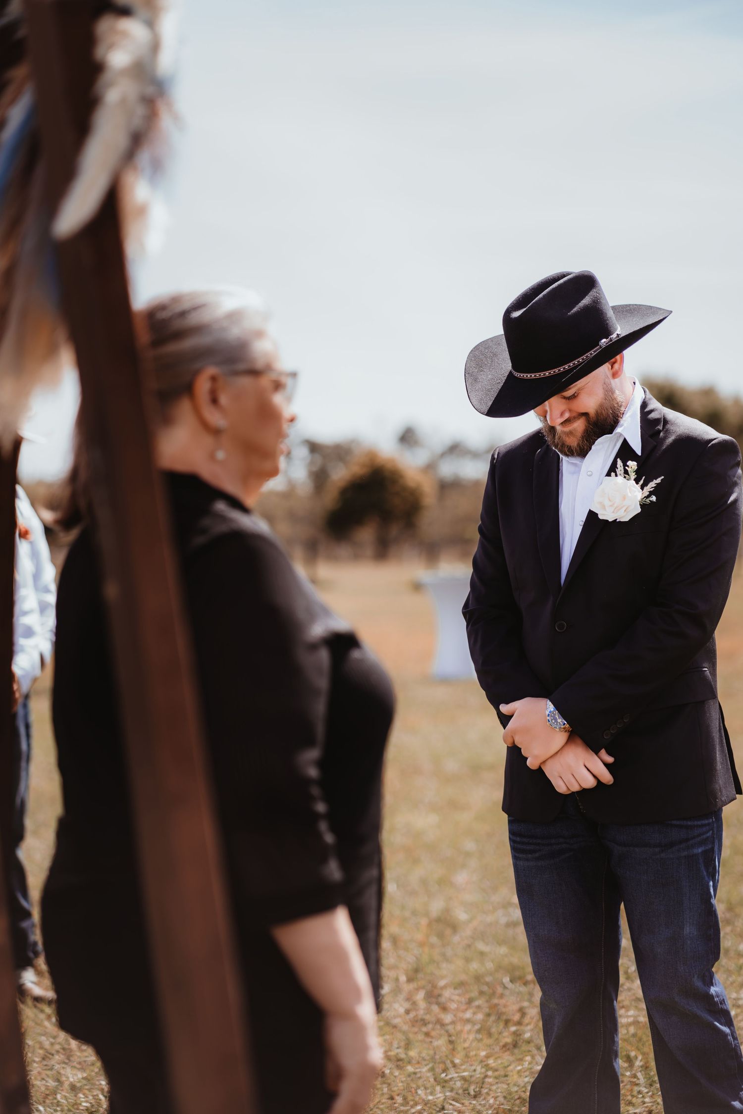 Groom in black cowboy hat and tuxedo jacket with white boutonniere looks down during outdoor wedding ceremony.