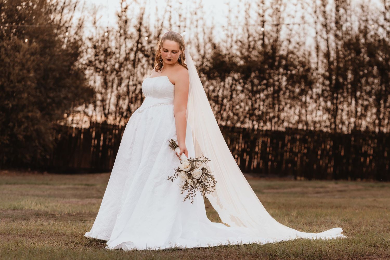 Elegant bride in white strapless gown holds bouquet while posing in golden evening light.
