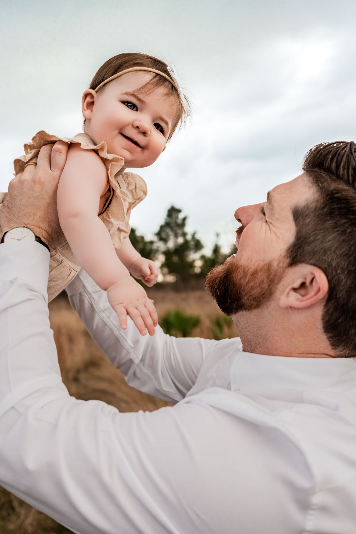 Bearded father in white shirt lifting smiling baby girl in tan dress against cloudy sky outdoors.