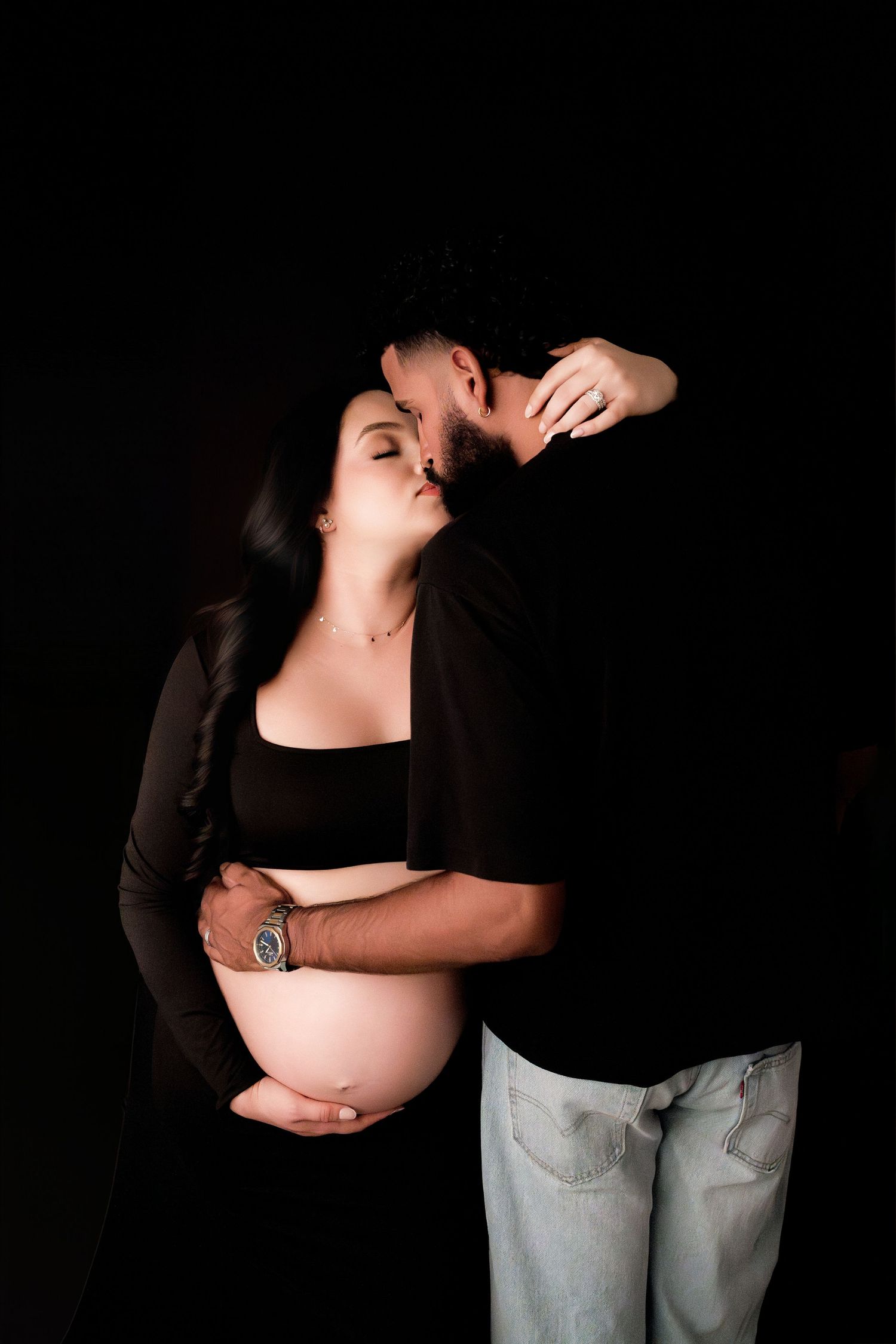 Expecting couple sharing a kiss during maternity session, man embracing woman's pregnant belly in black outfits.