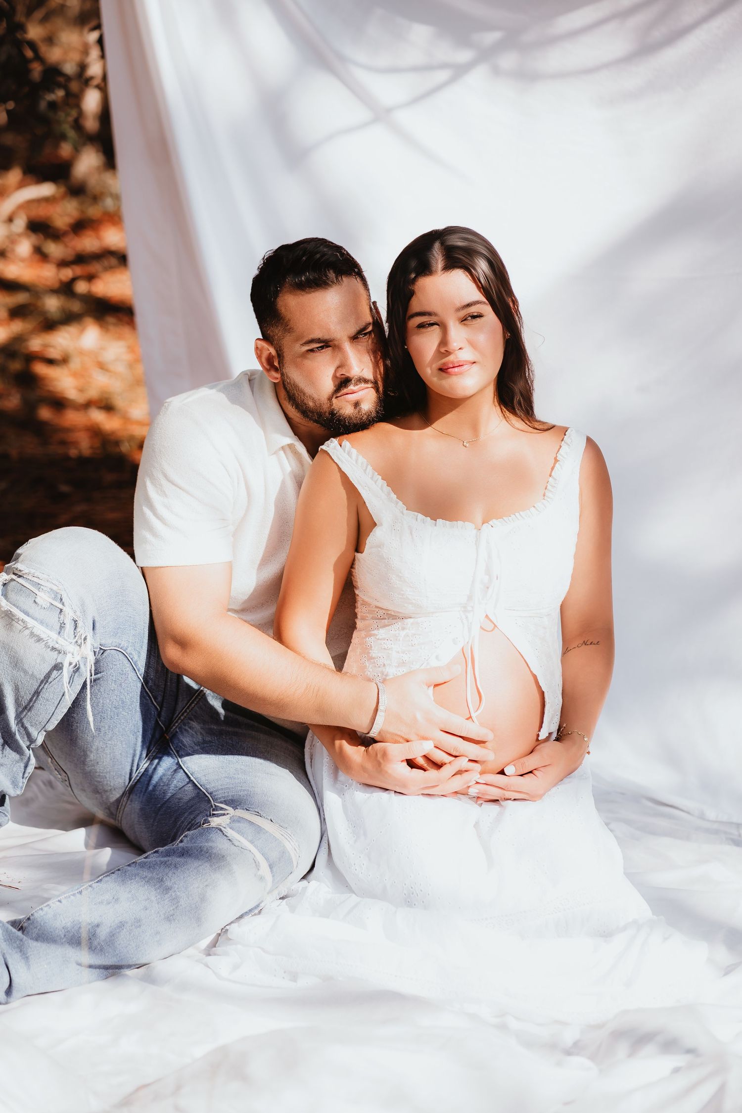 Couple sitting together with man embracing pregnant woman in white dress on white fabric.