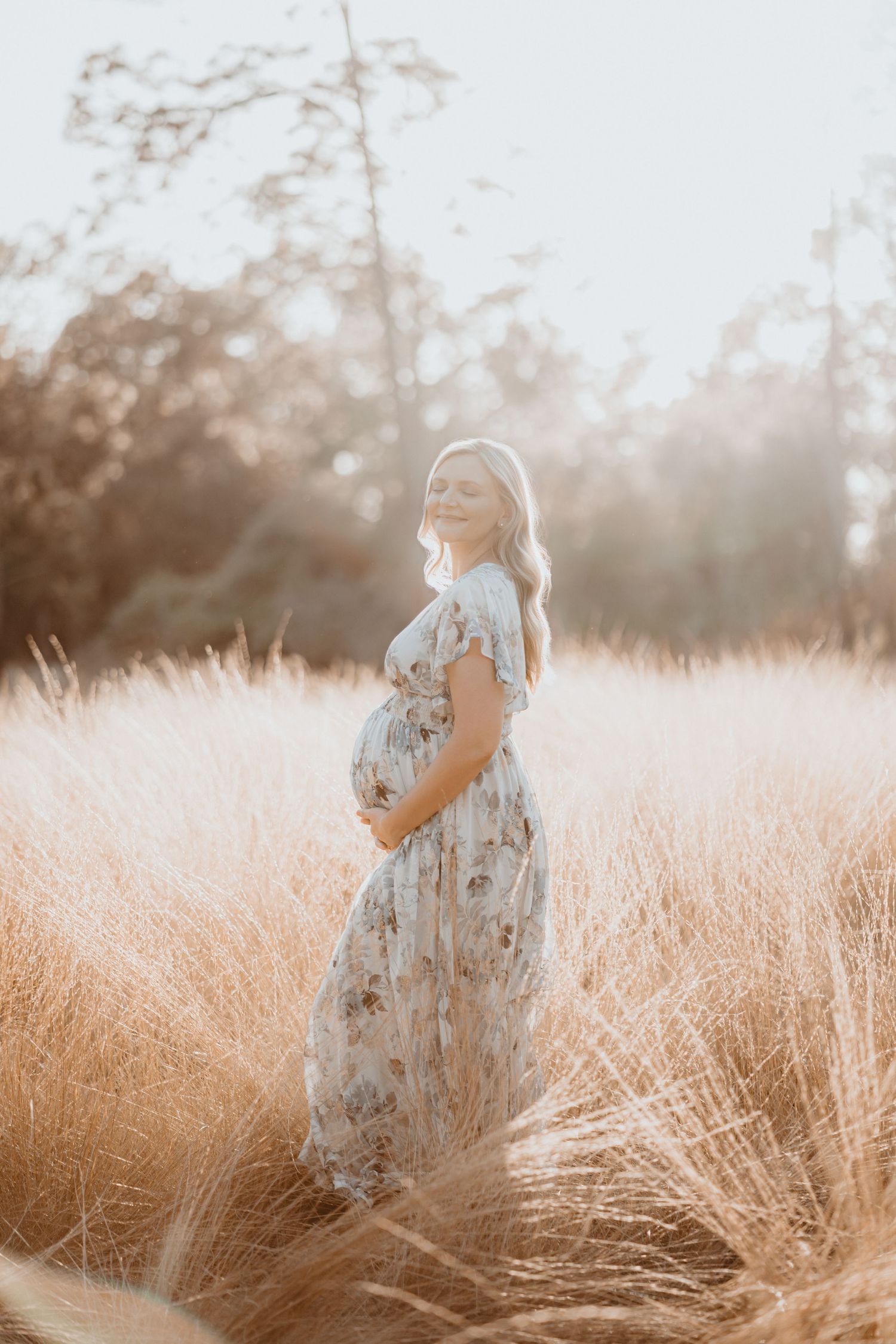 Glowing pregnant woman in floral dress cradles baby bump while standing in a sunlit golden grass field.