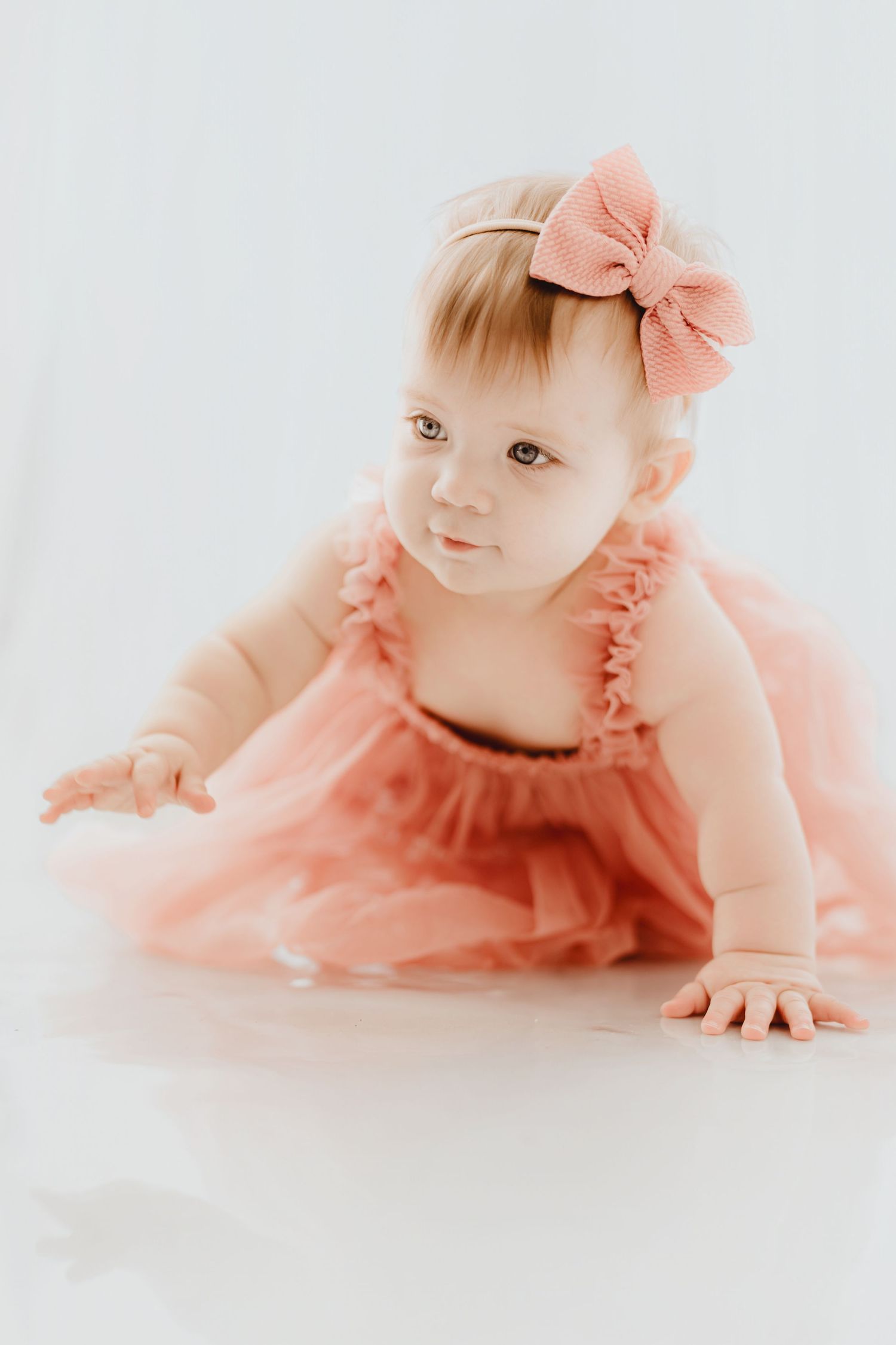 Baby girl in pink tulle ruffle dress and bow headband crawling on white floor.