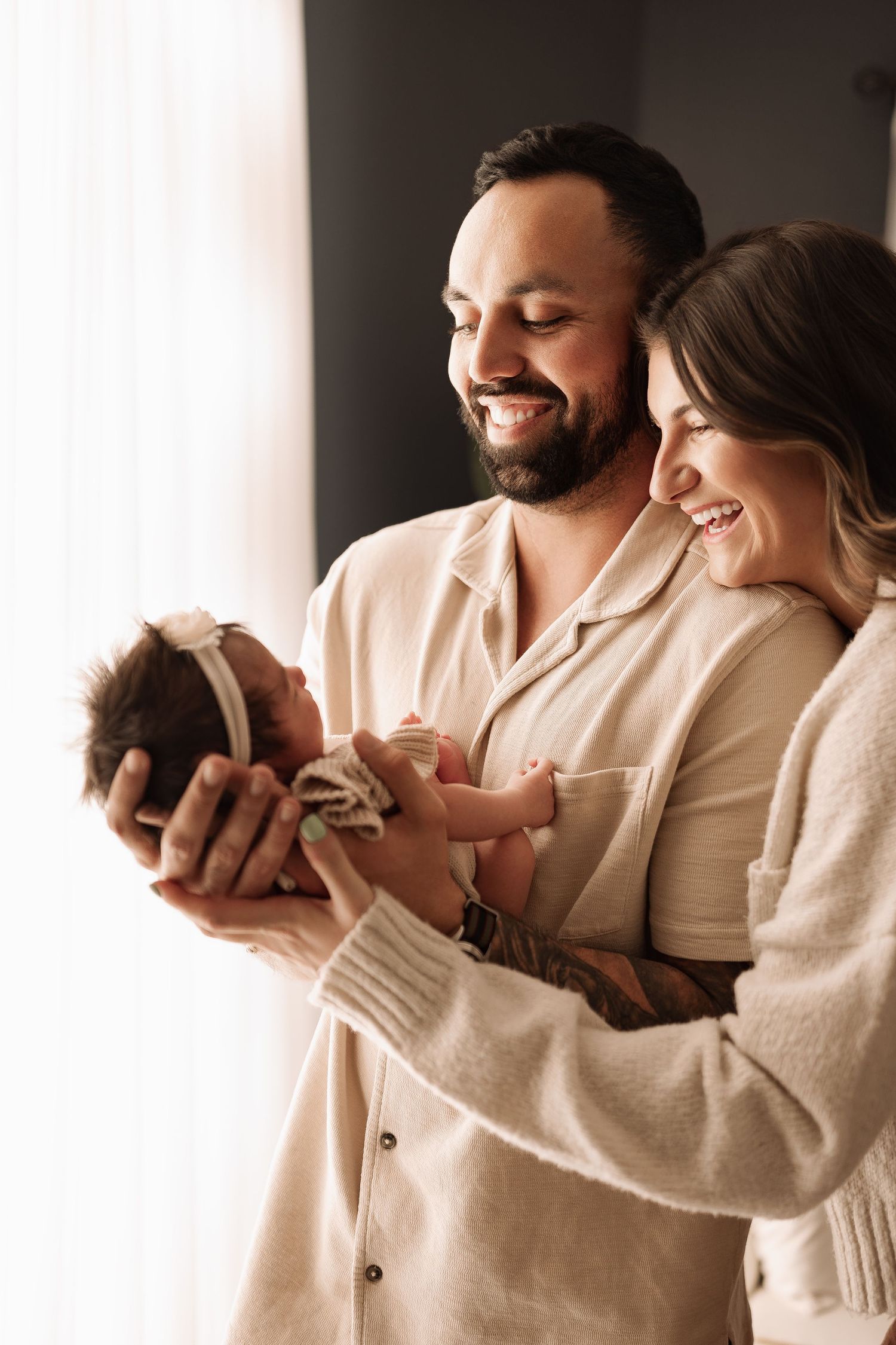 A laughing couple shares a joyful moment while holding their newborn baby girl by a window.