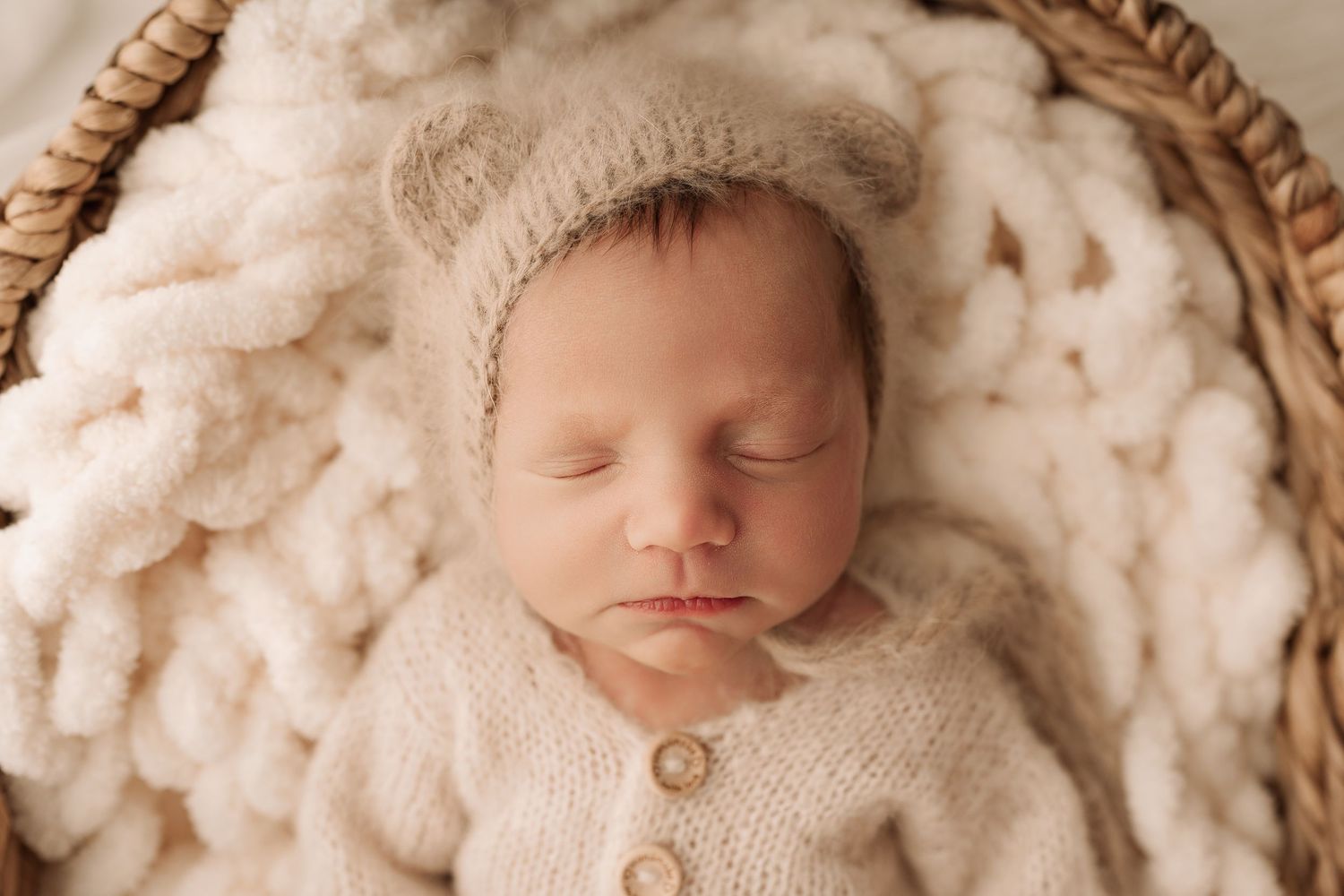 Sleeping newborn baby in a bear ear bonnet nestled in a wicker basket with white blanket.