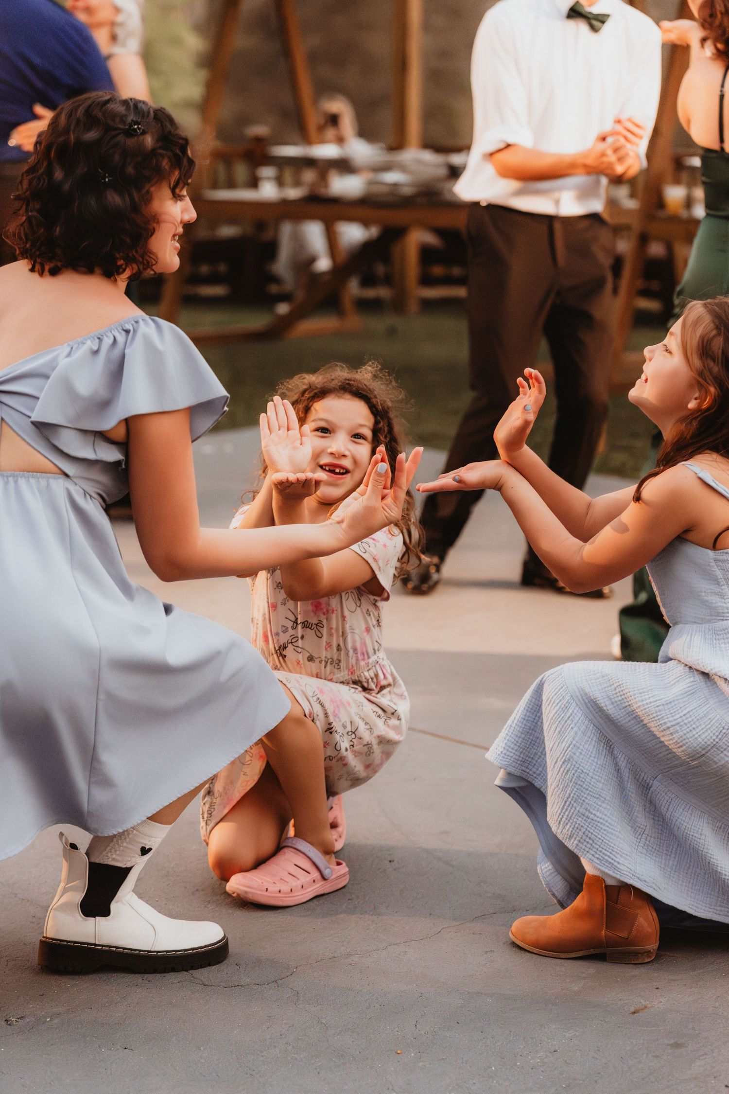 Two bridesmaids in blue dresses crouching to play with a smiling young child on dance floor.