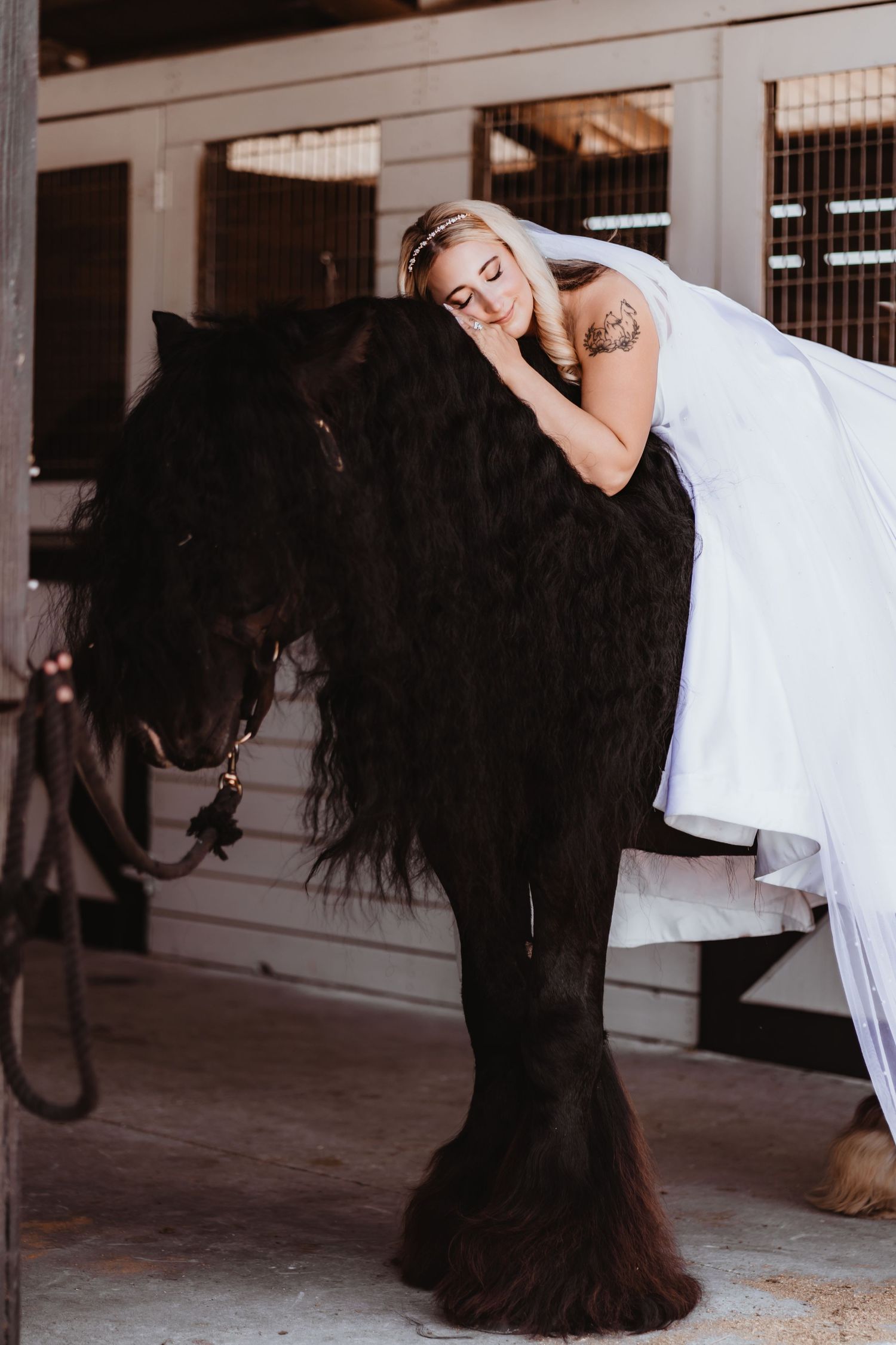 Bride in white wedding dress leans lovingly on a black horse in a stable.