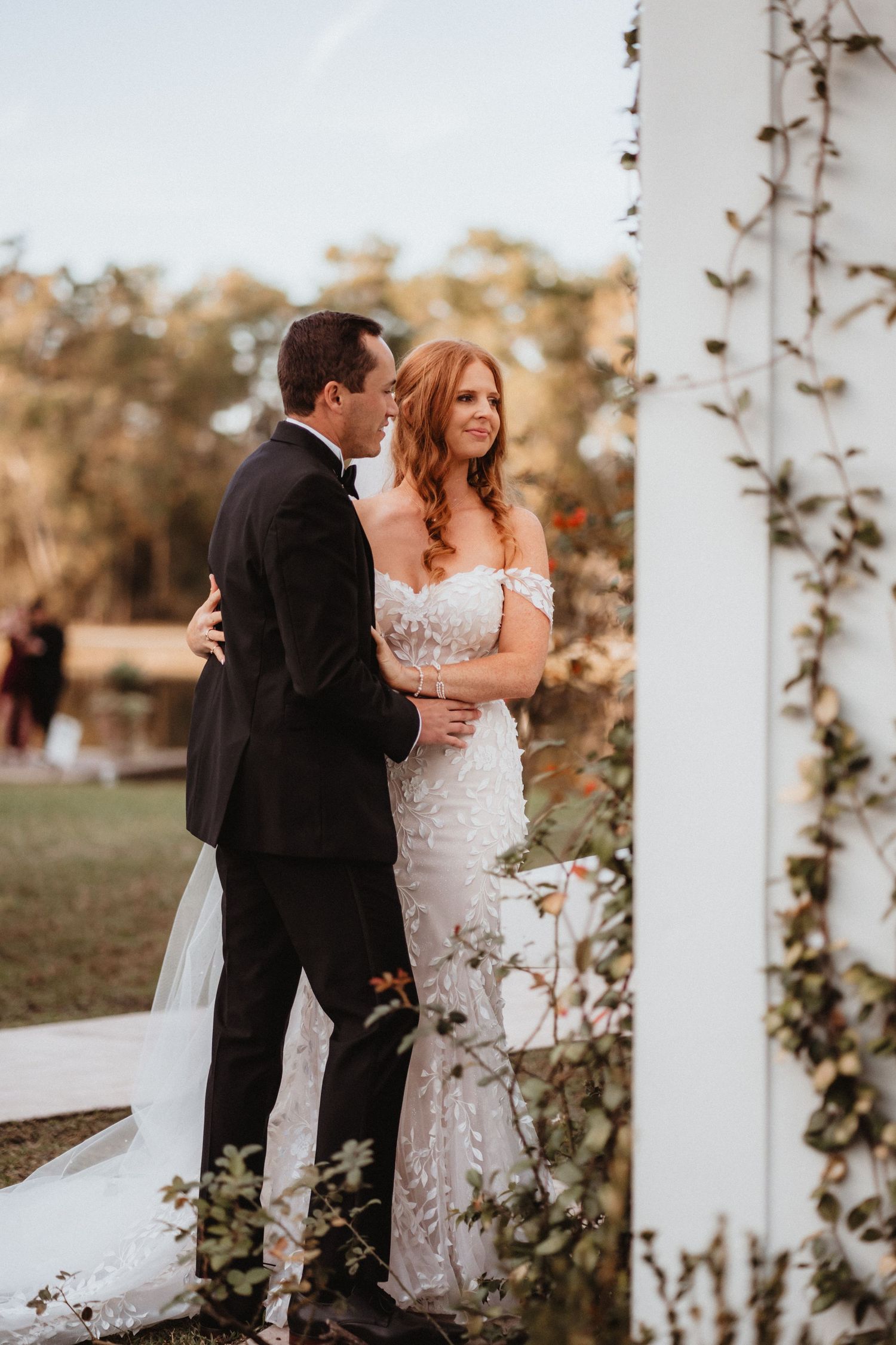Bride in lace gown and groom in tuxedo share a romantic embrace near a white garden arbor.