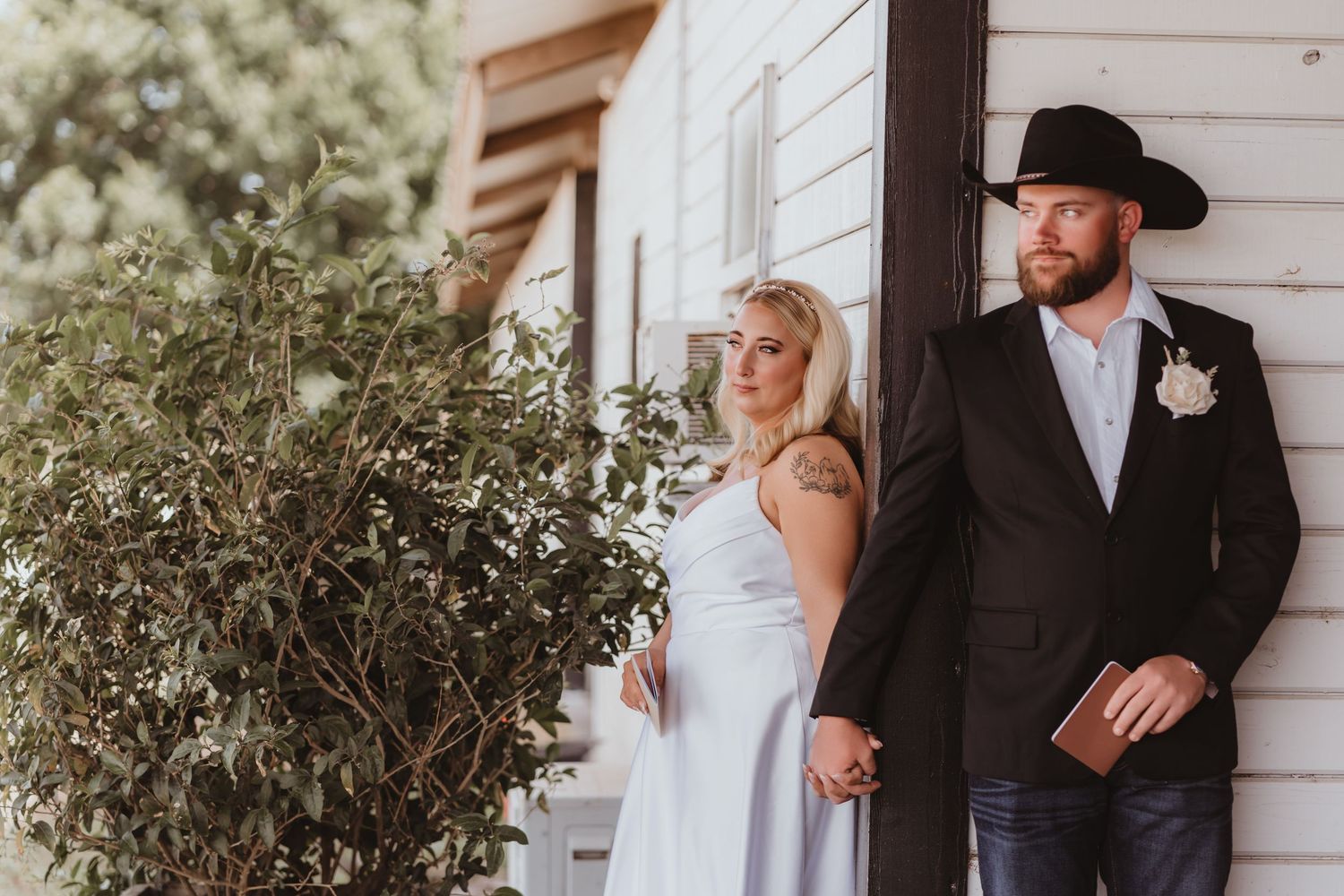 Bride in white gown and groom in black cowboy hat hold hands beside lush greenery at farmhouse.