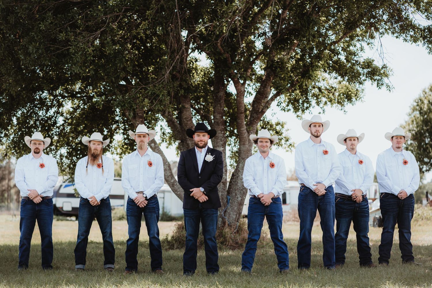Groom in black suit flanked by eight groomsmen in white shirts and cowboy hats outdoors.