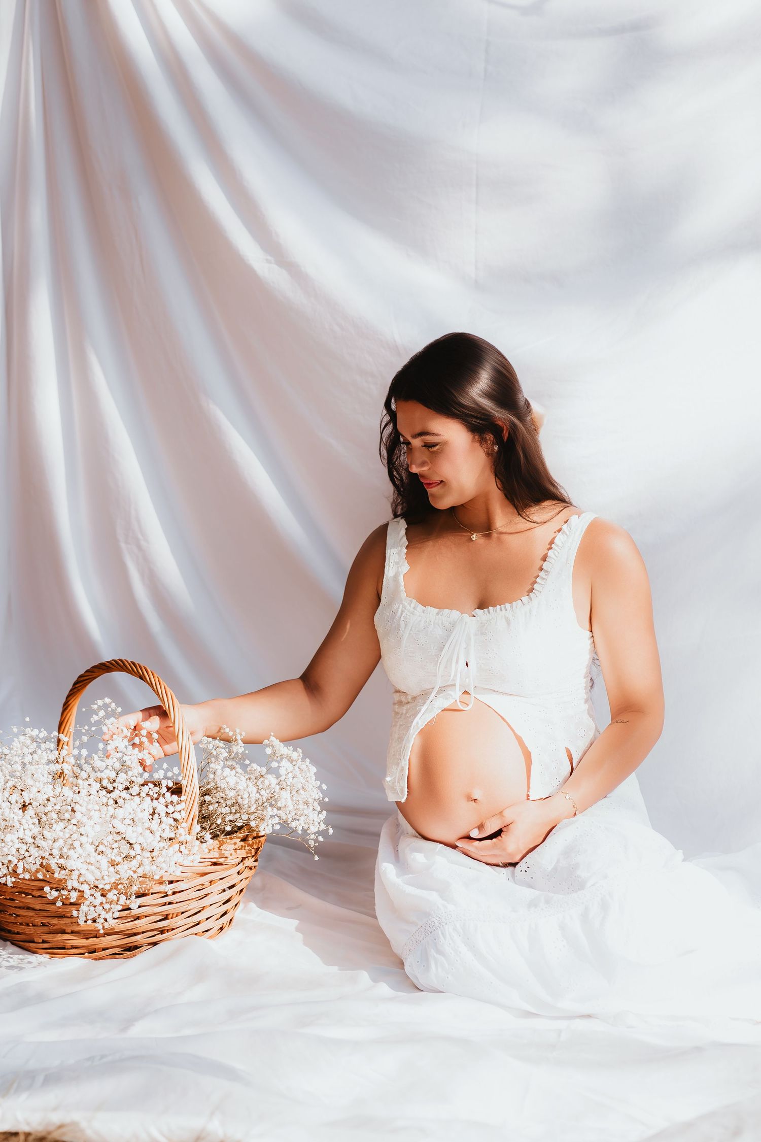 Pregnant woman in white dress sitting beside wicker basket of white flowers on fabric backdrop.