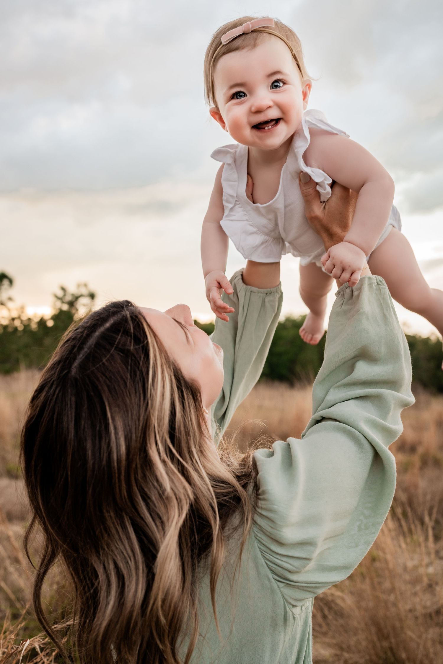 Joyful baby girl giggling as mother lifts her high in an outdoor golden grass field at sunset.