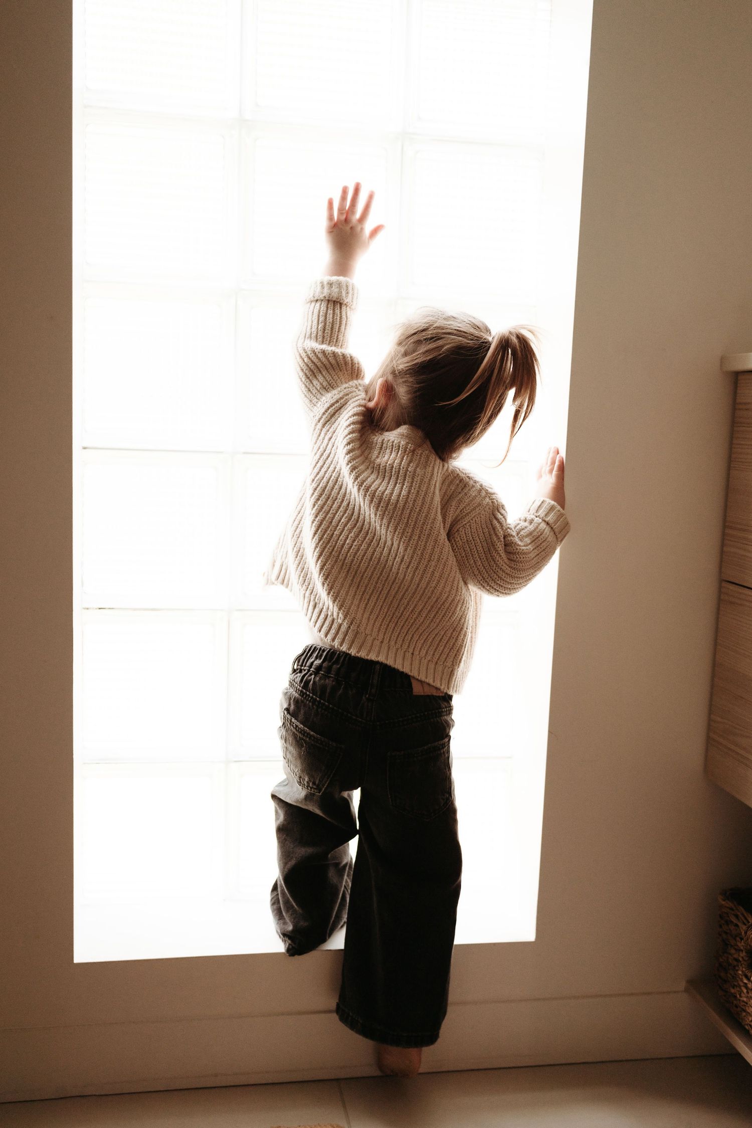 A young girl in a beige sweater and jeans reaches up toward a bright sunlit window.