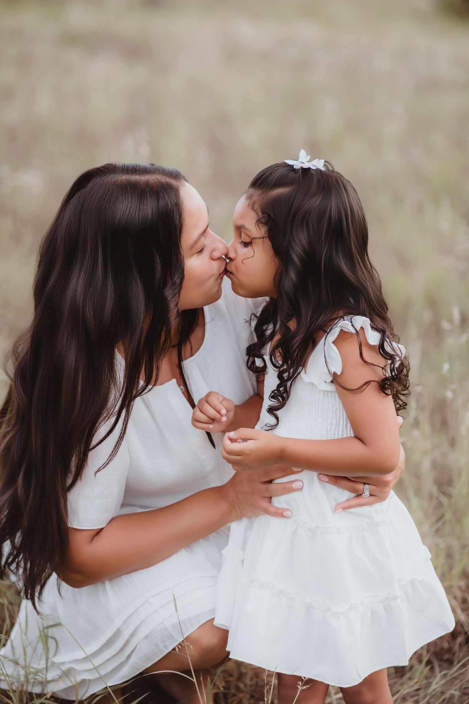 Mother and young daughter sharing a kiss while sitting in a grassy field outdoors.