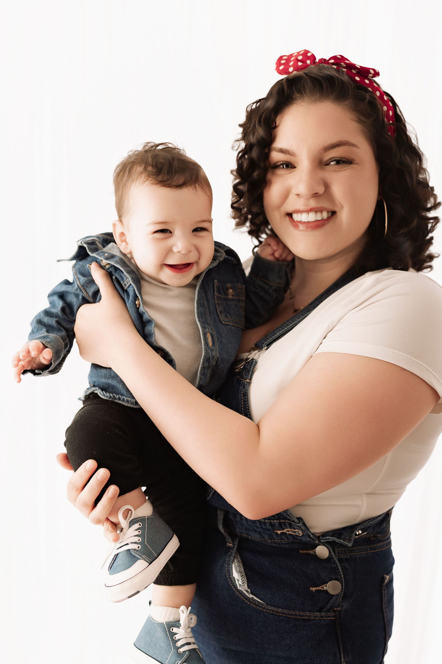Smiling curly-haired mother in denim overalls holding her laughing baby dressed in a denim jacket.