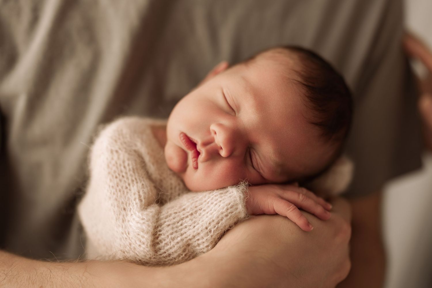 Newborn baby sleeping peacefully in parent's hands wrapped in a cream knit blanket.
