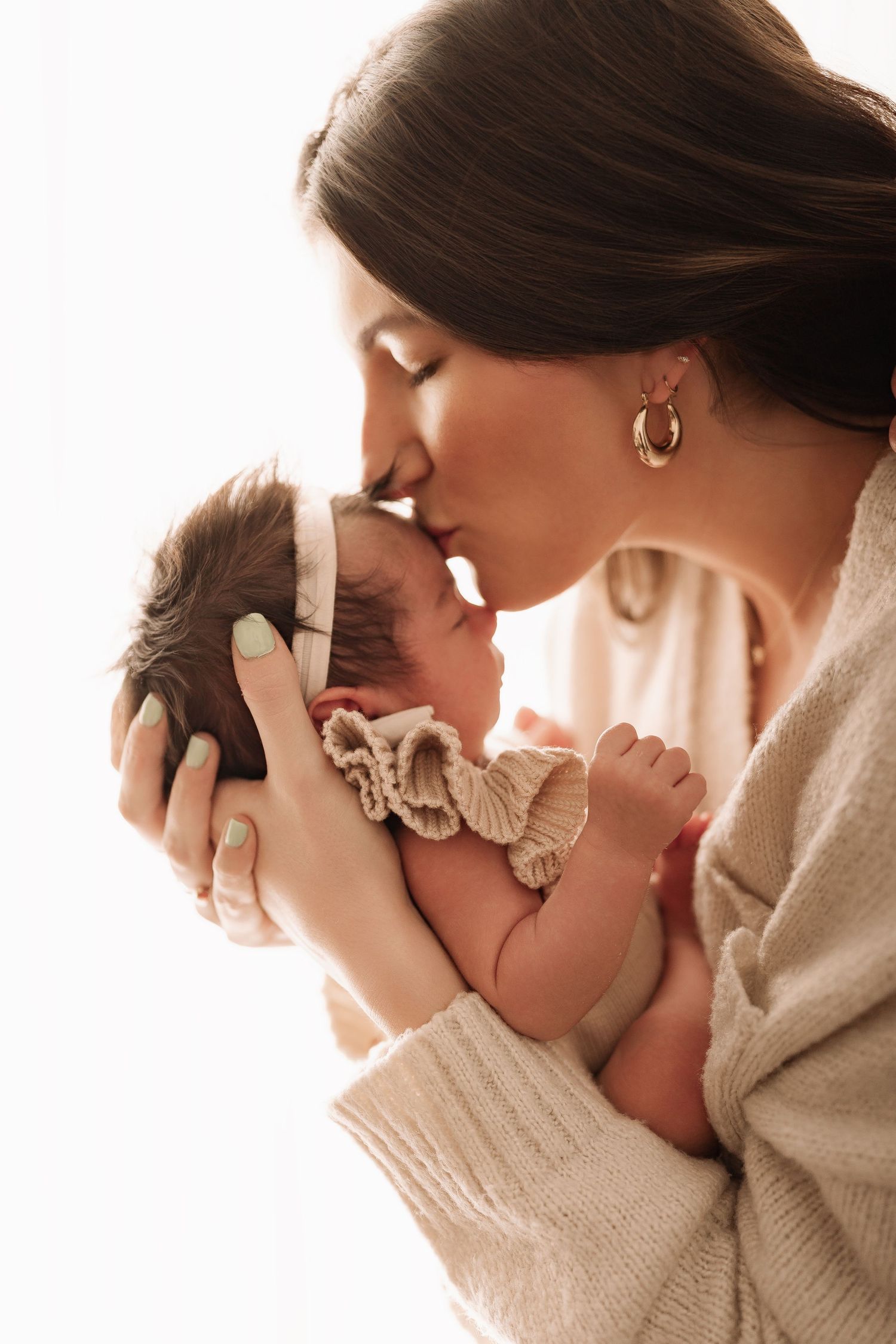 A mother tenderly kisses her newborn baby girl while holding her gently in both hands.