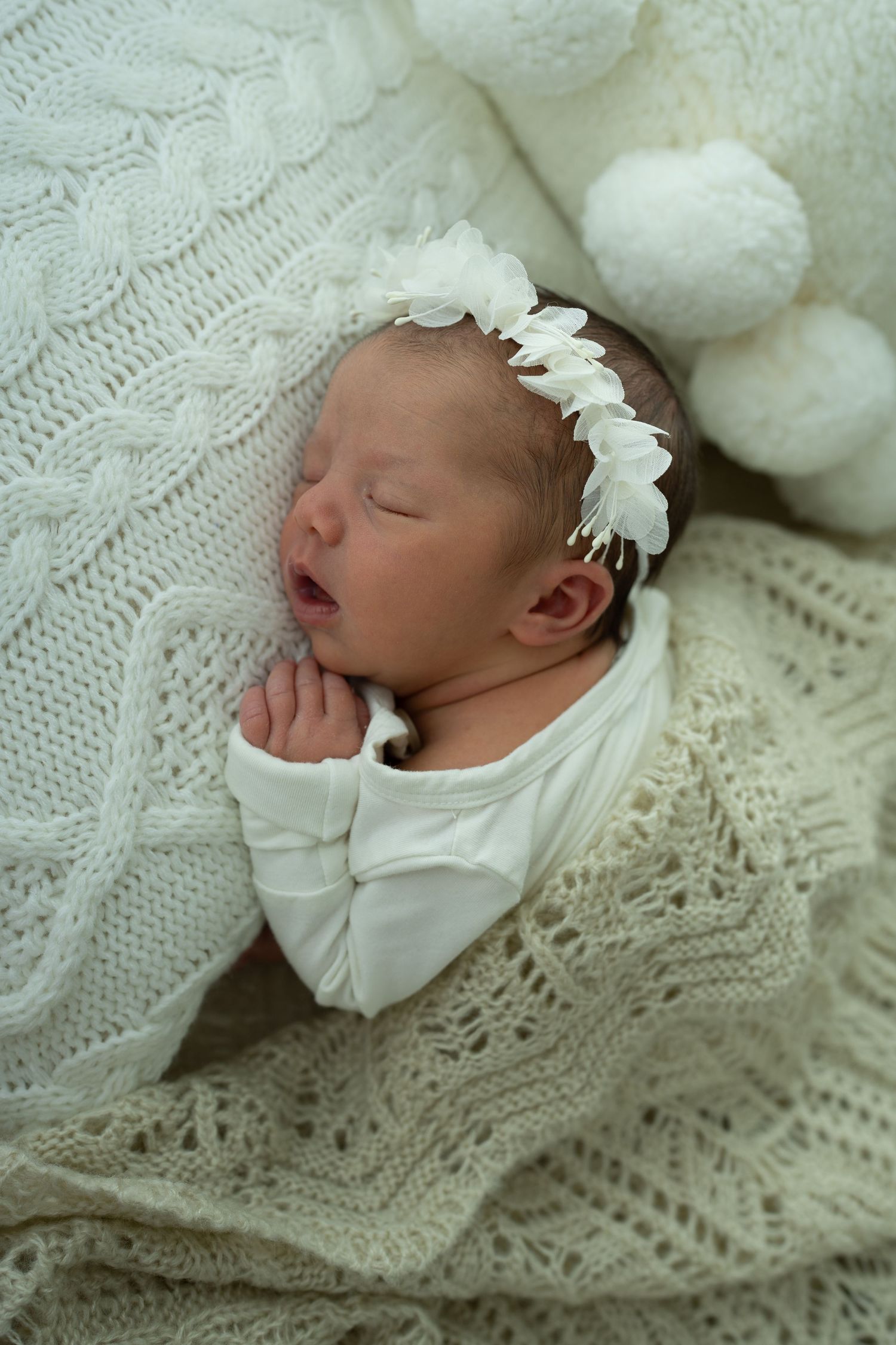 Sleeping newborn baby girl in white outfit and floral headband, wrapped in cream knit blanket with pom pom decor.