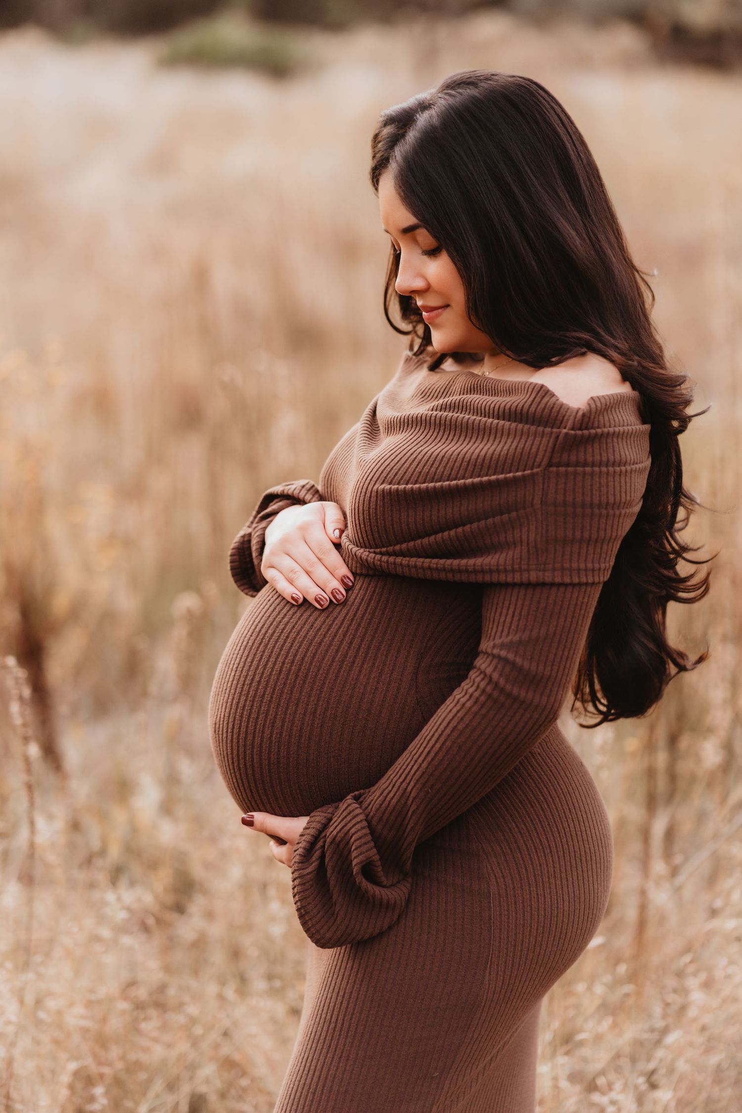 Pregnant woman in brown ribbed off-shoulder dress cradling baby bump in golden field during maternity photoshoot.