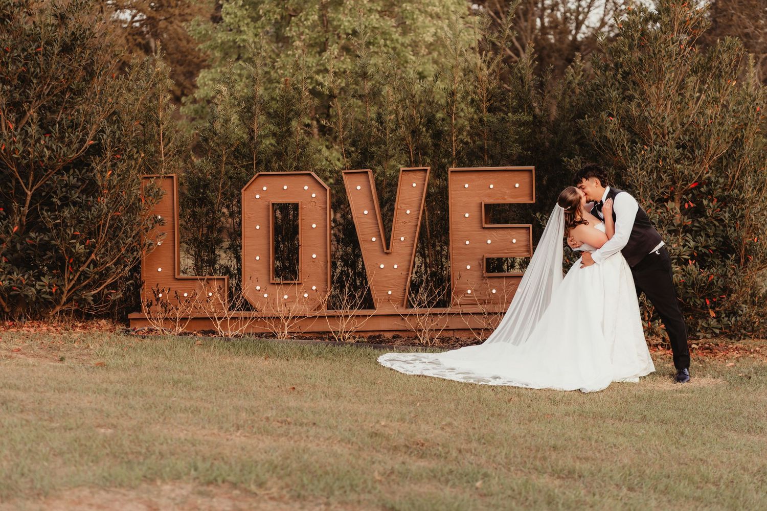 Bride and groom sharing a kiss in front of a rustic illuminated LOVE marquee sign outdoors.