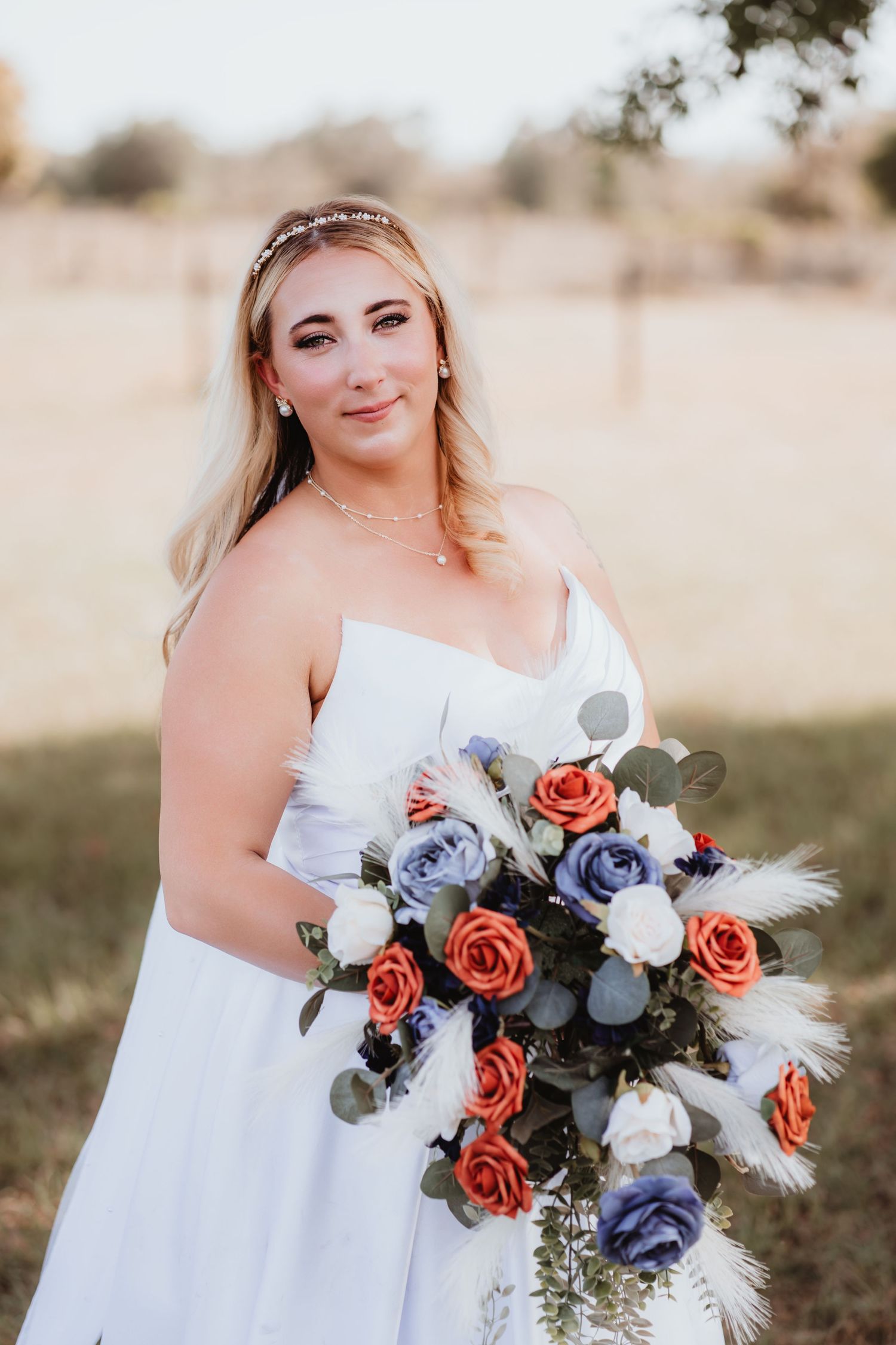 Blonde bride in strapless white gown holding cascading bouquet of orange, blue, and white roses.