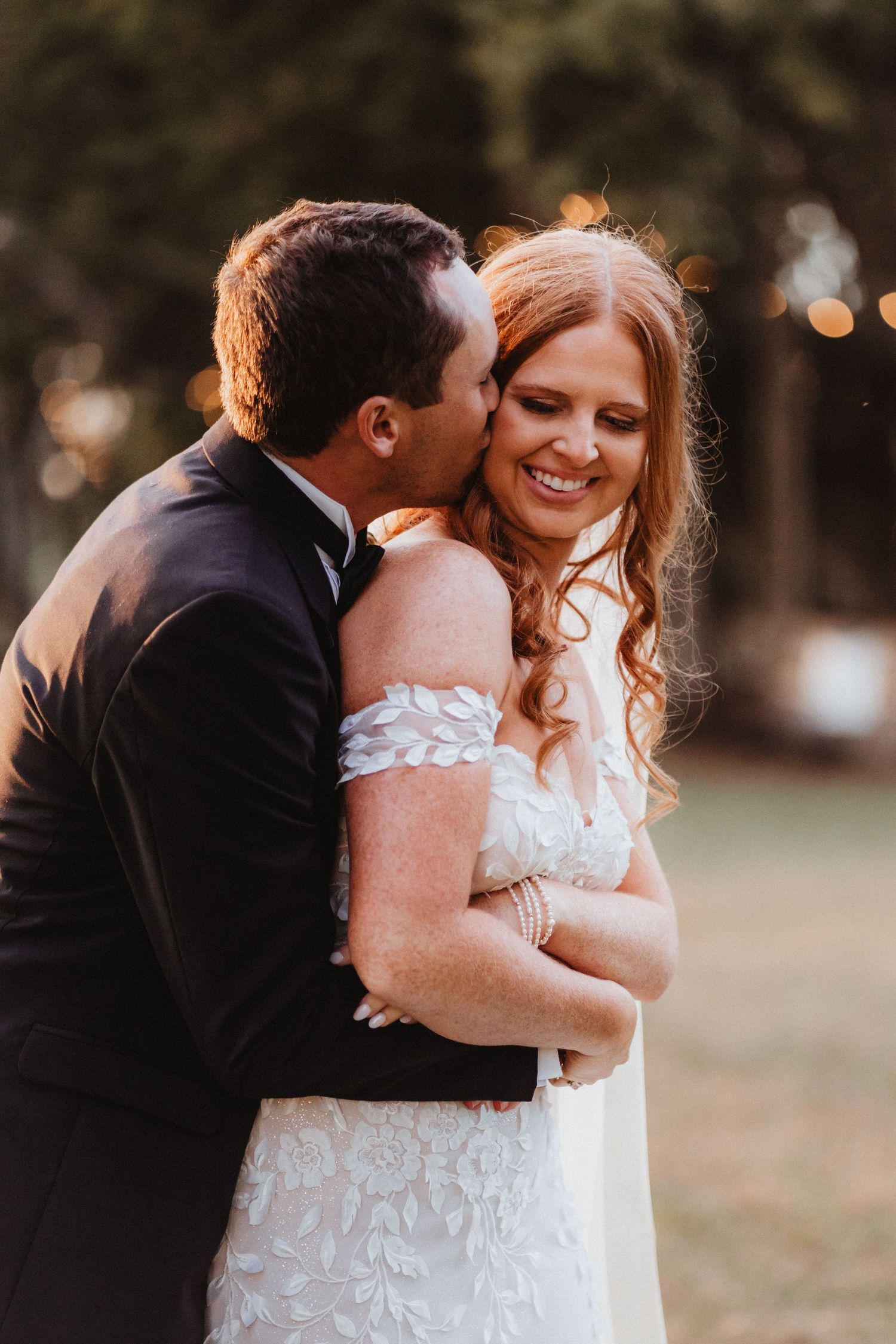 Groom in black tuxedo kisses smiling bride on cheek, bride in off-shoulder lace gown with floral appliques.
