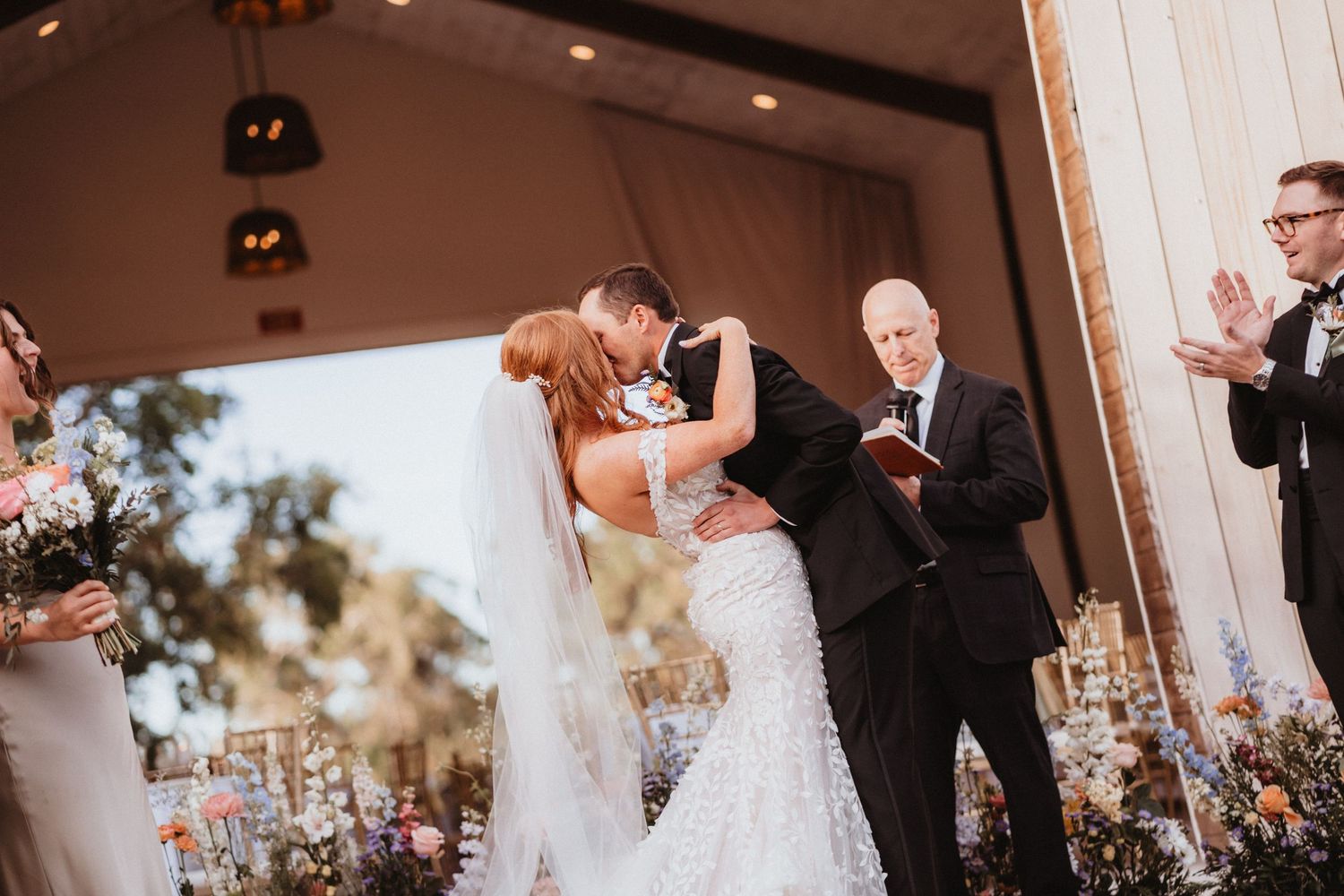 Bride and groom share first kiss at outdoor wedding ceremony while officiant and guests look on.
