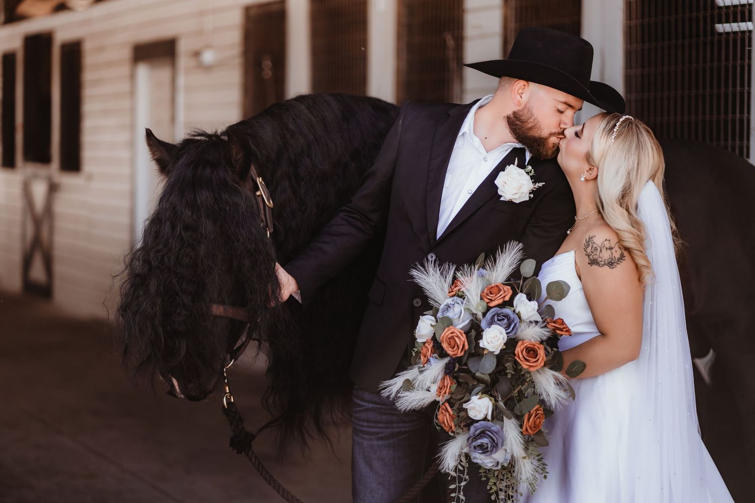 Couple in western wedding attire kiss near a black horse outside stable.