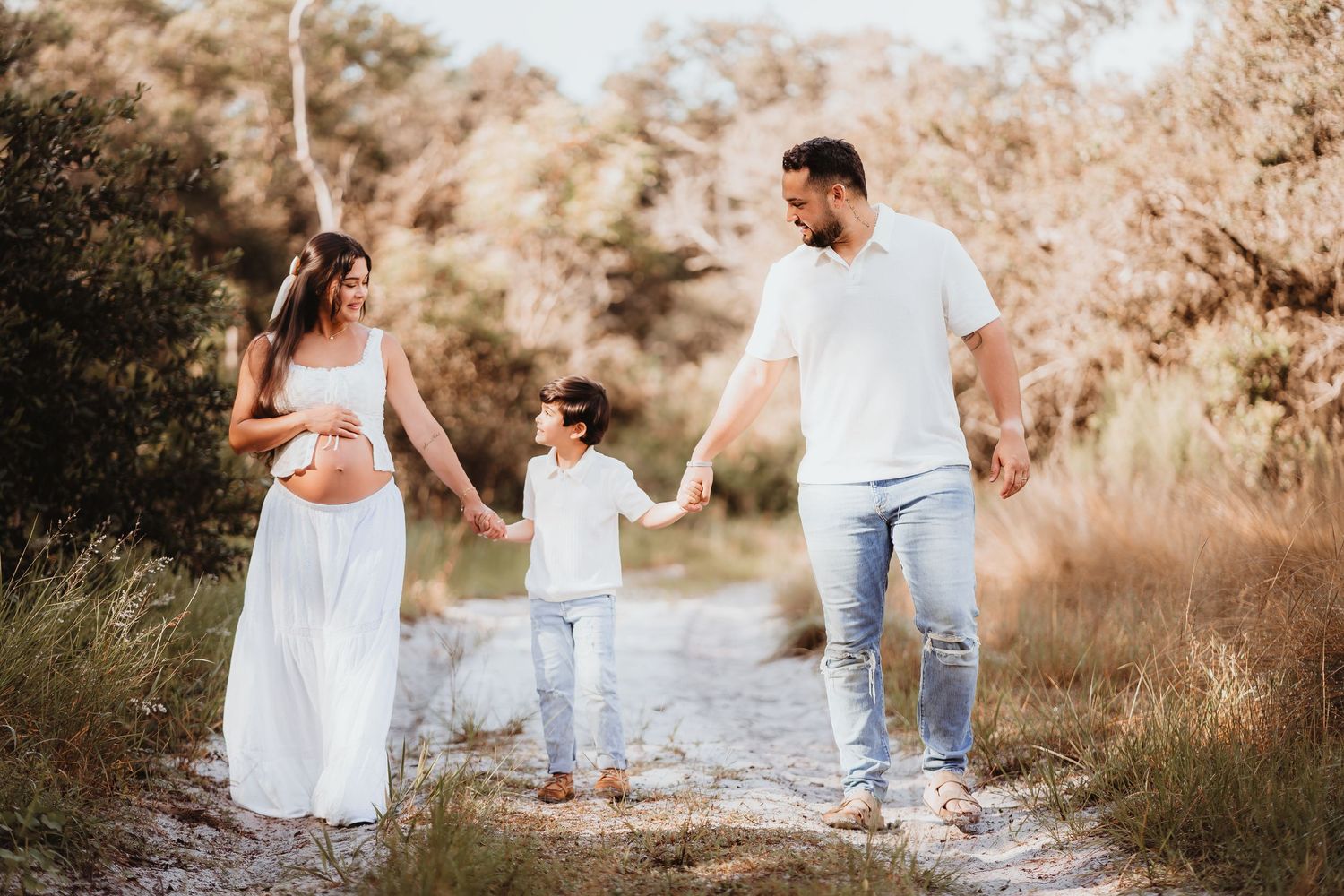 Expecting family in white outfits holding hands with young child on sunlit outdoor nature trail.