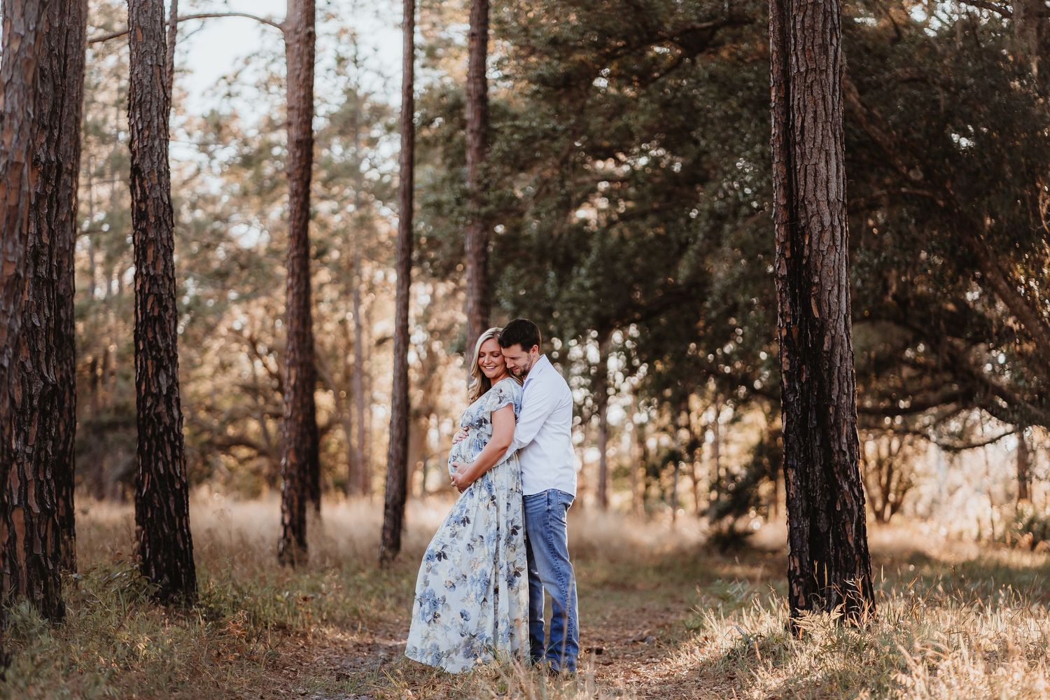 Couple embracing tenderly in a sunlit pine forest during maternity photoshoot.