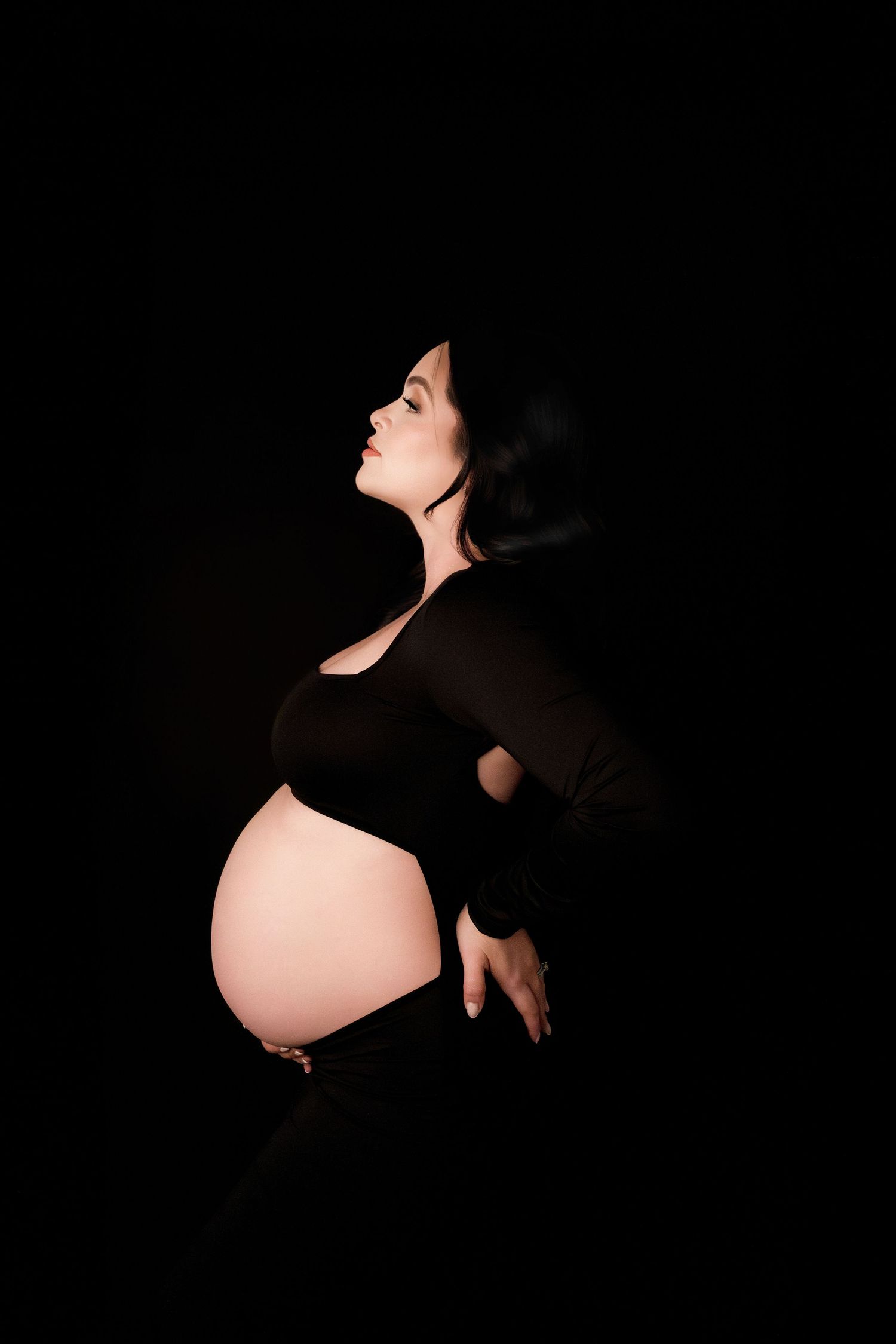 Elegant side profile of pregnant woman in black outfit against dark background.