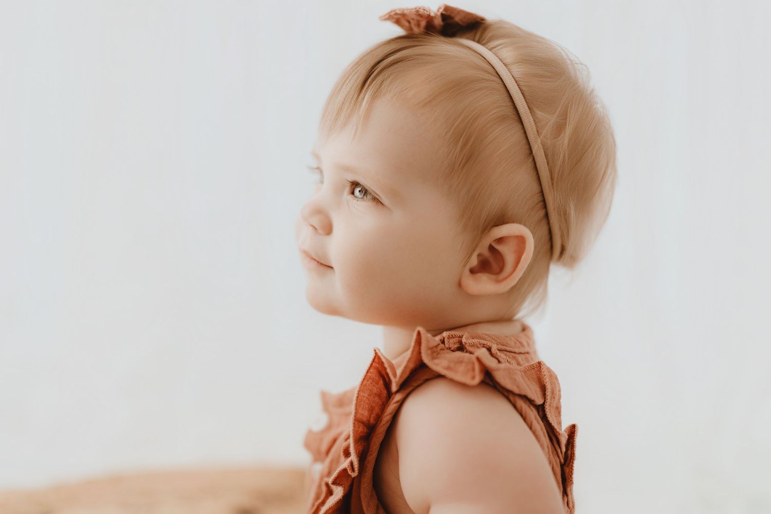 Side profile of cute baby girl wearing rust romper and bow headband in studio.