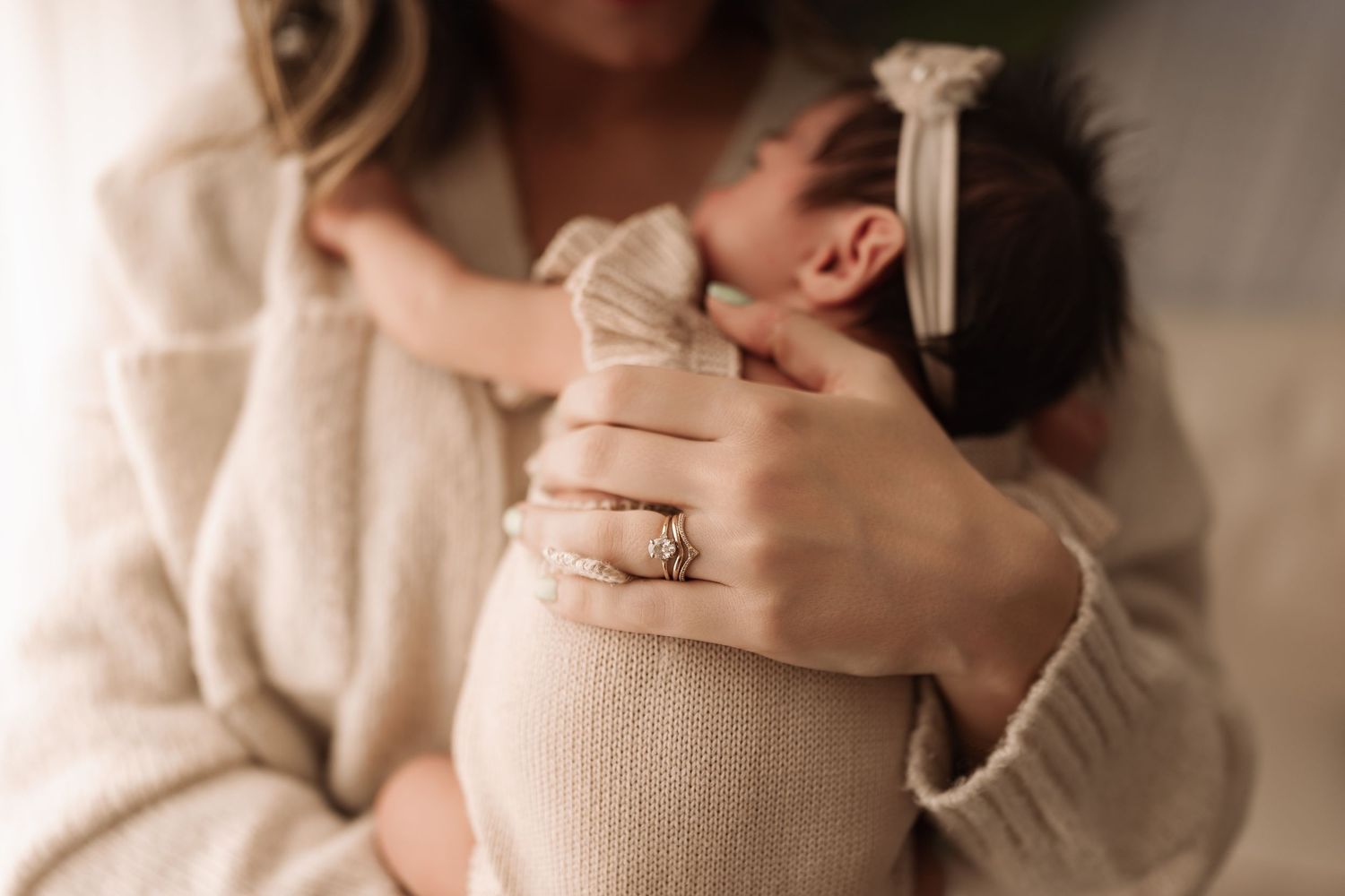 Close-up of mother's hands holding swaddled newborn baby with white bow headband, ring visible.