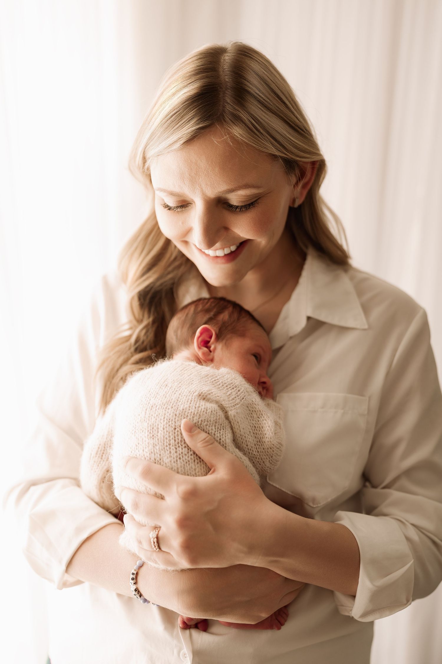Blonde mother in white shirt smiling down at her newborn baby wrapped in a cream knit blanket.