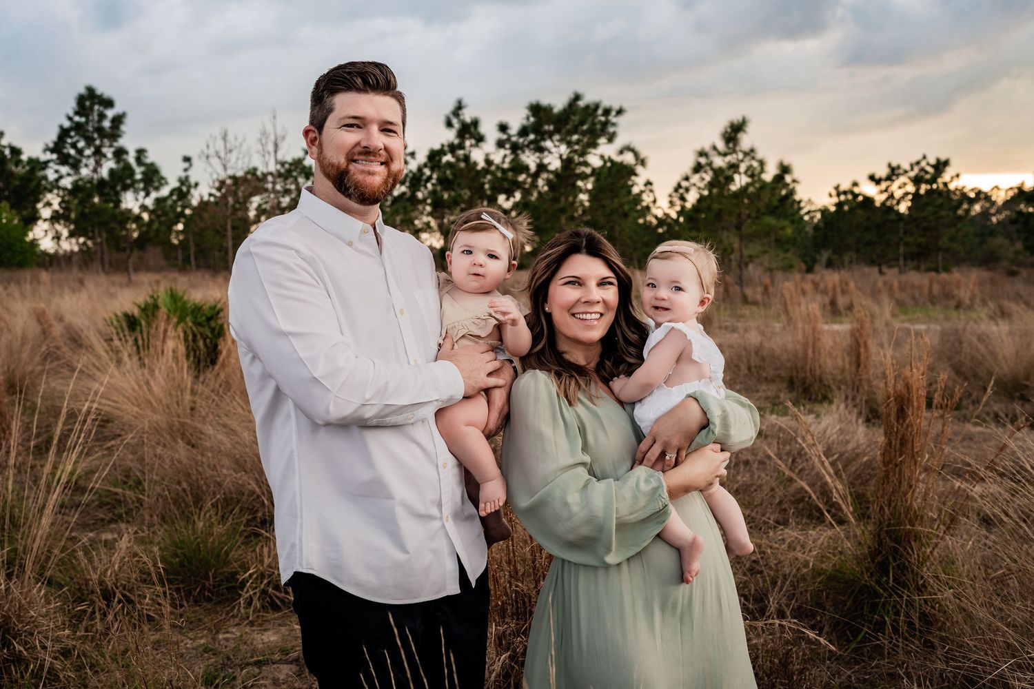 Smiling family of four holding two babies in a golden grass field during sunset outdoors.