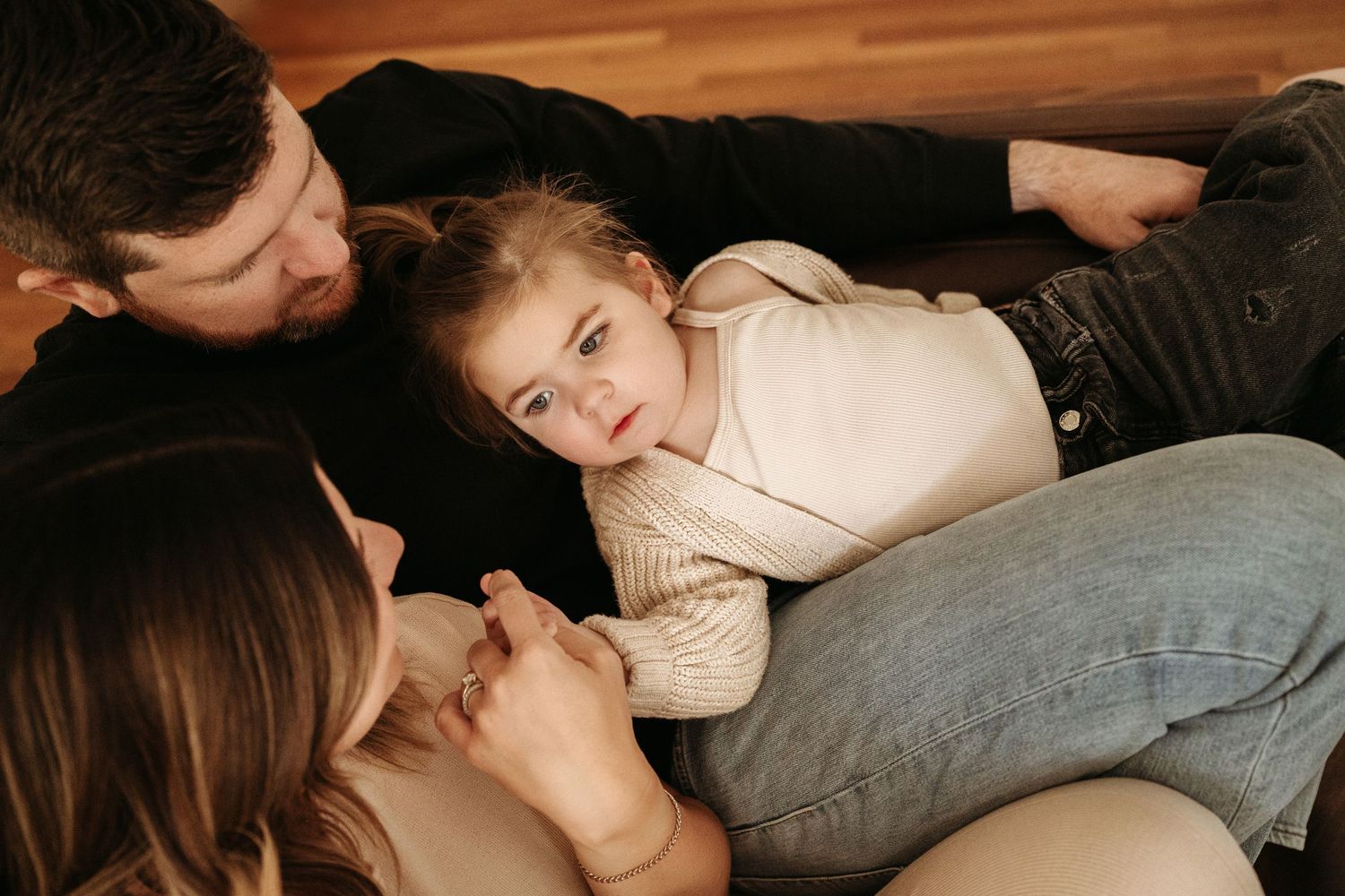 A young child lies between parents on a wooden floor, looking up with curious eyes.