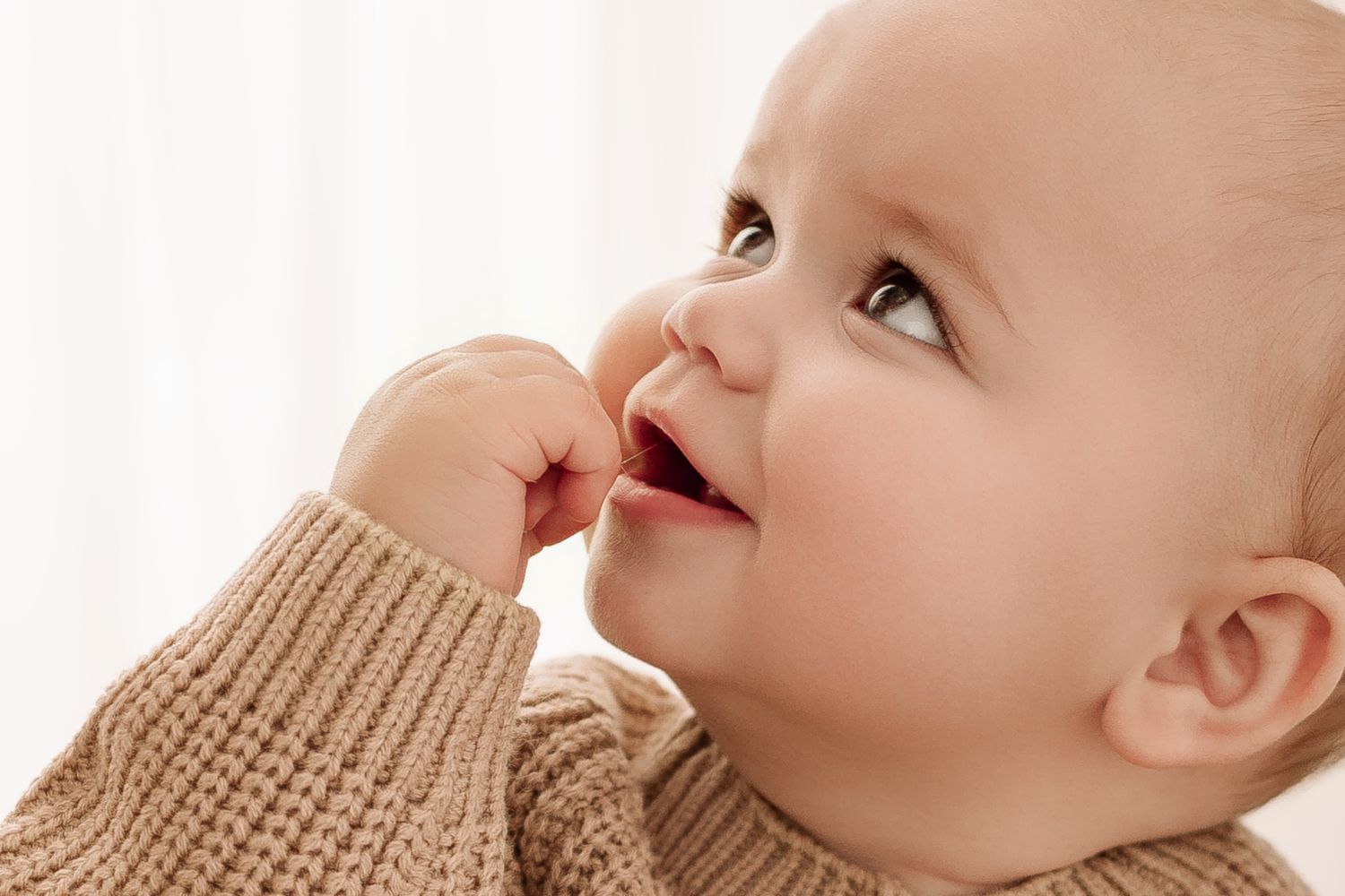 Adorable baby in a beige knit sweater looking up curiously with finger near mouth.