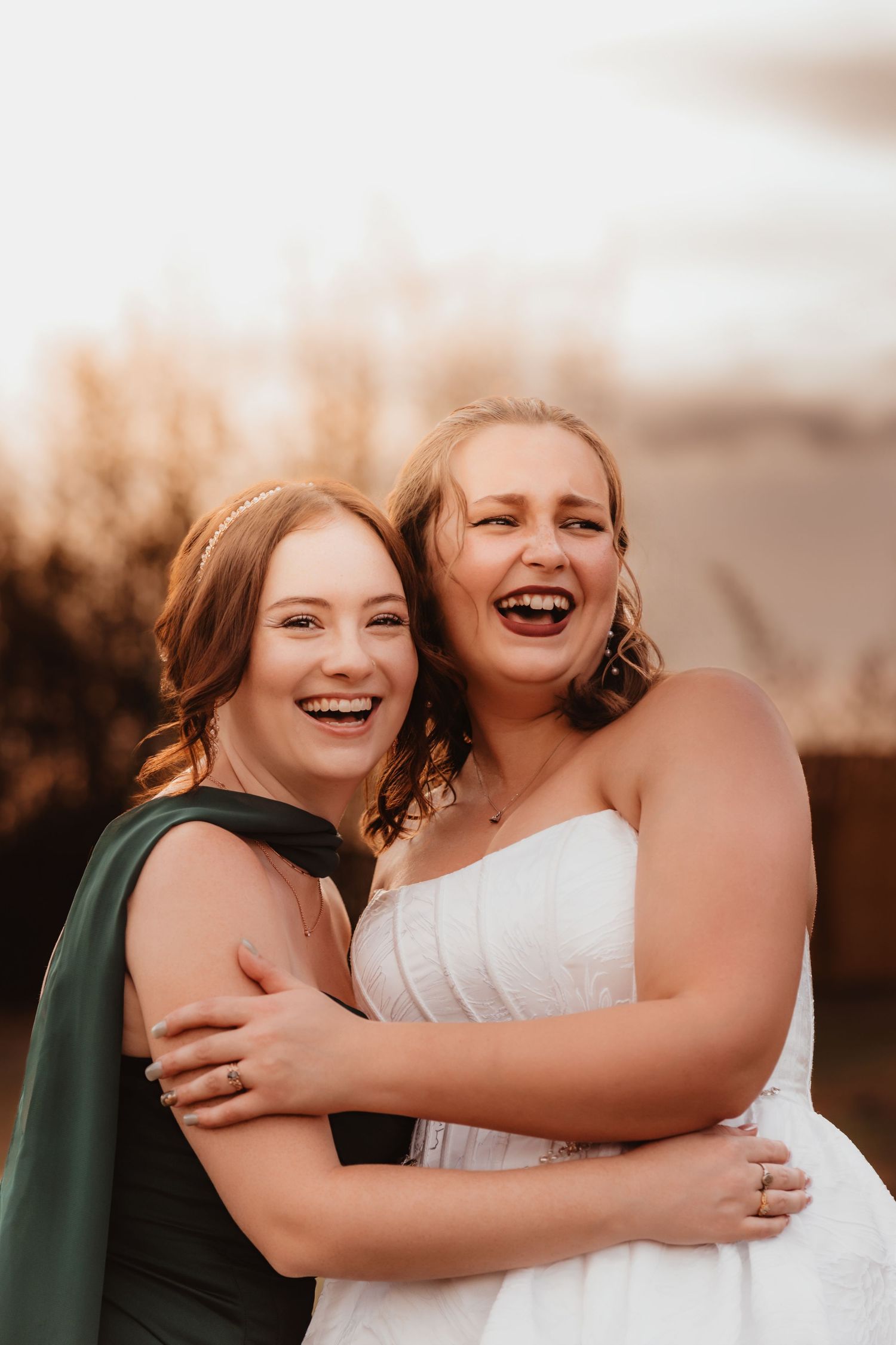 Bride in white strapless gown and bridesmaid in green dress laugh and embrace outdoors.