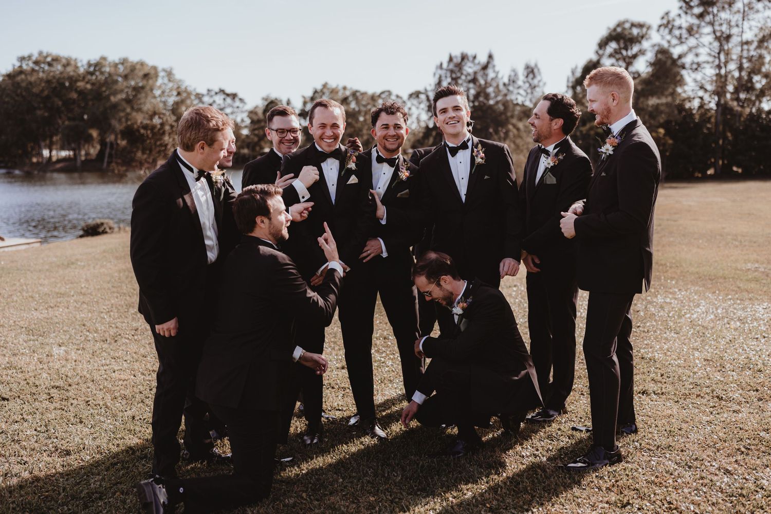 Groomsmen in black tuxedos laughing and celebrating together outdoors near a lake on a wedding day.