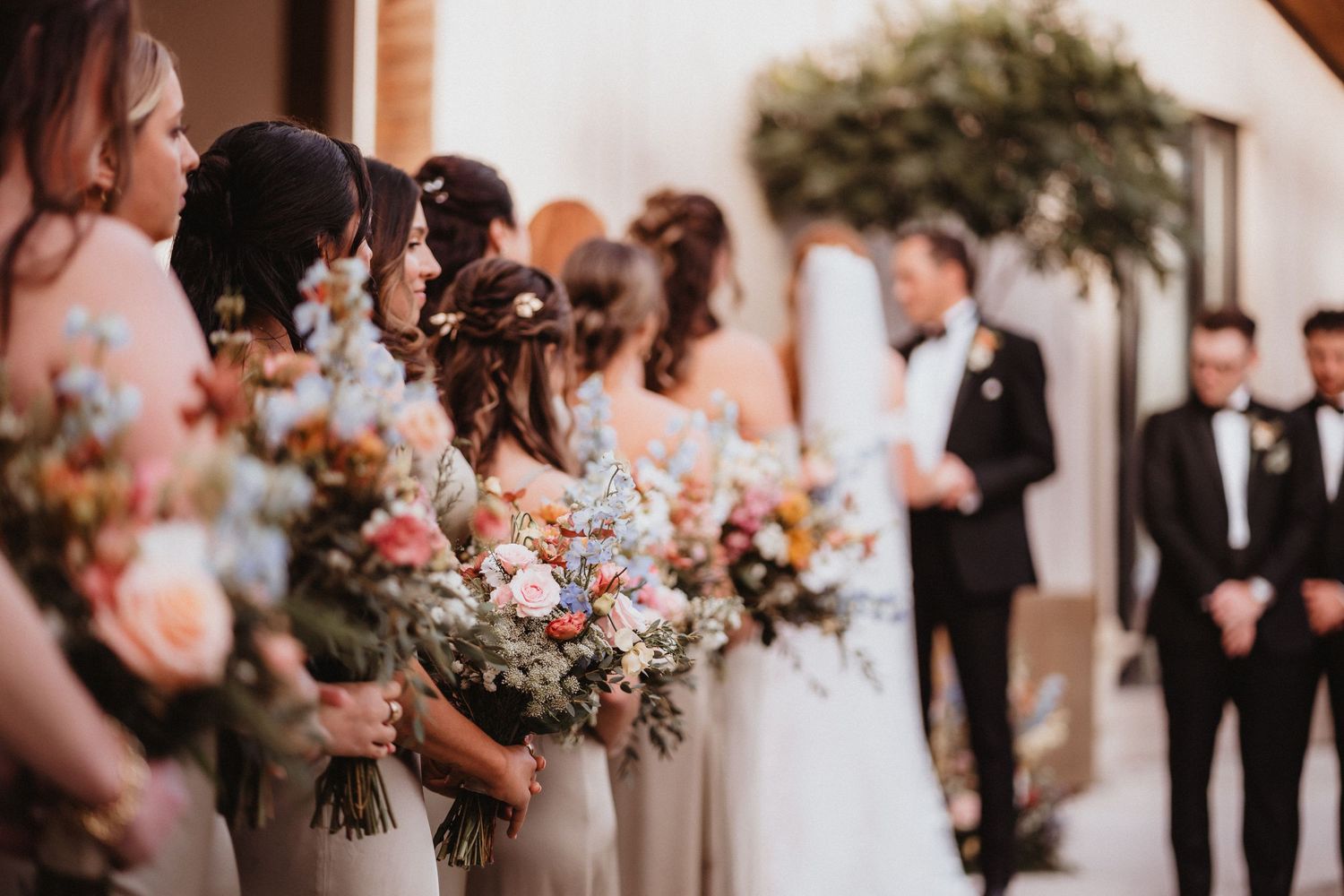 Bridesmaids in champagne gowns holding colorful floral bouquets stand at outdoor wedding ceremony.