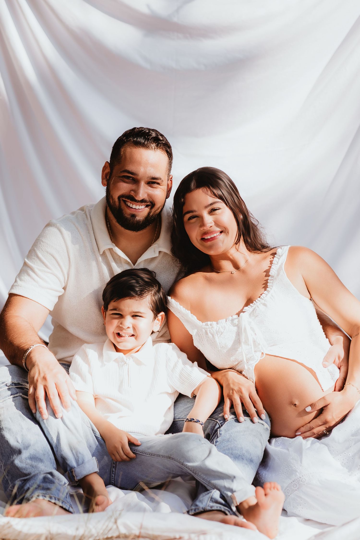 Happy family with pregnant mother, father, and young son posing together in white outfits.