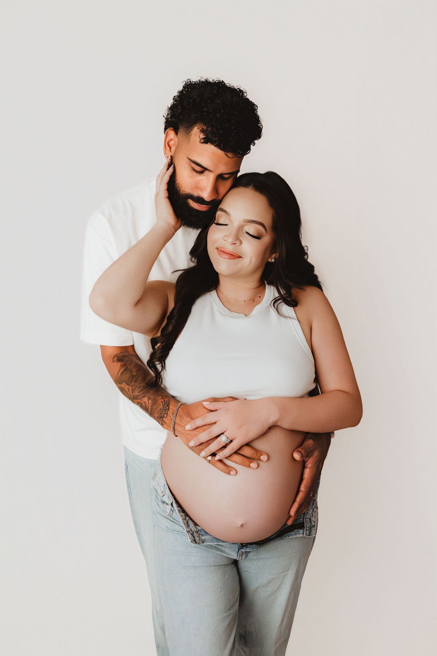Couple embracing during maternity session, man with tattoos hugging pregnant woman in white crop top.
