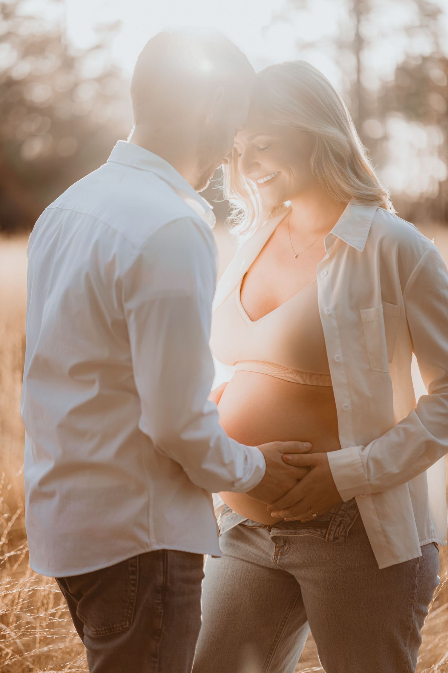 Smiling pregnant woman and partner embracing in sunlit field of tall grass.