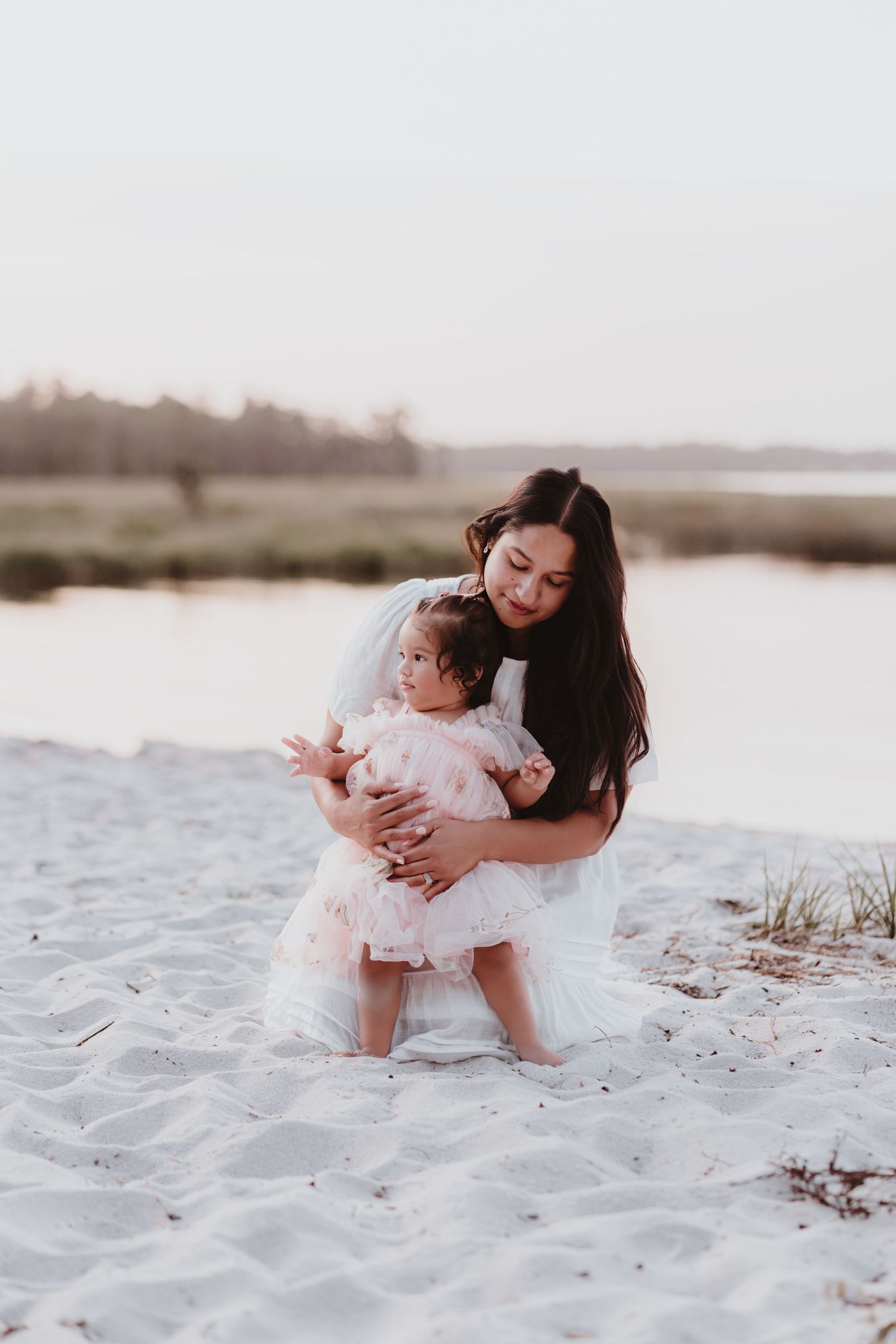 Mother in white dress kneeling on sand cuddling baby girl in pink dress.