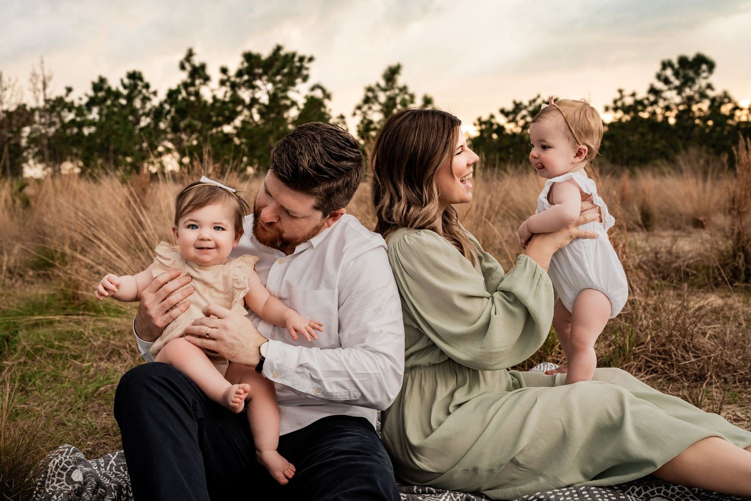 Parents sitting back to back on a blanket each holding a baby girl in a sunny outdoor field setting.