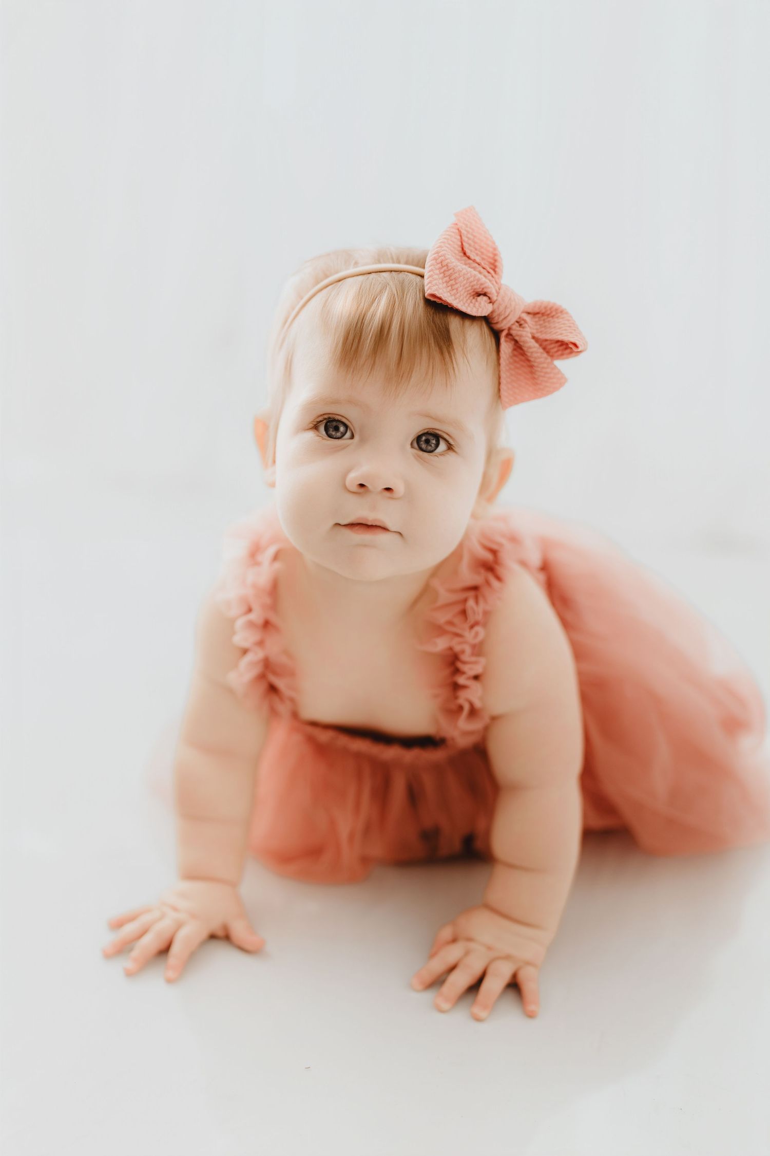 Baby girl in coral ruffle romper and pink bow headband gazing upward while crawling on white floor.