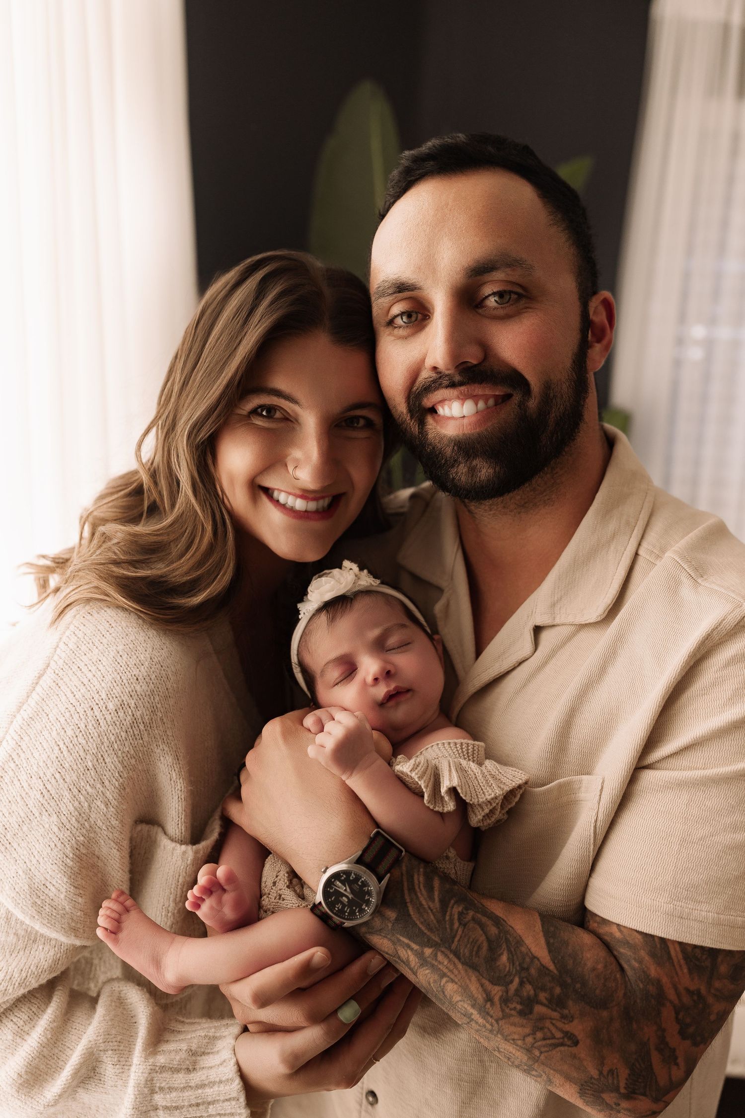 Smiling parents holding their sleeping newborn baby girl in a floral headband for a family portrait.